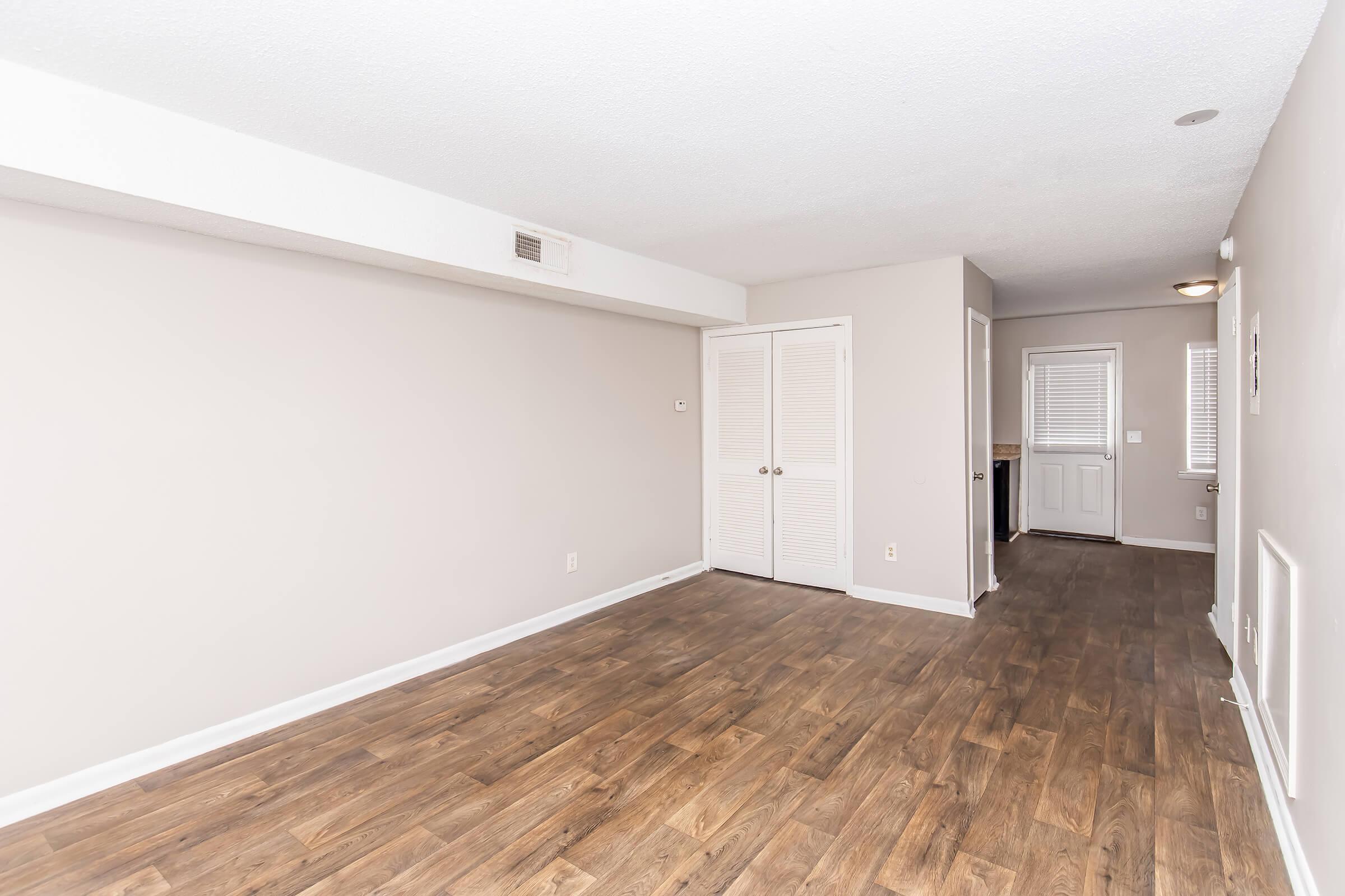 A spacious, empty living area with light-colored walls and a wooden floor. There are two doorways: one leading to a closet and the other to an exterior door. Natural light enters through a nearby window, creating a bright atmosphere. The overall look is modern and minimalistic.