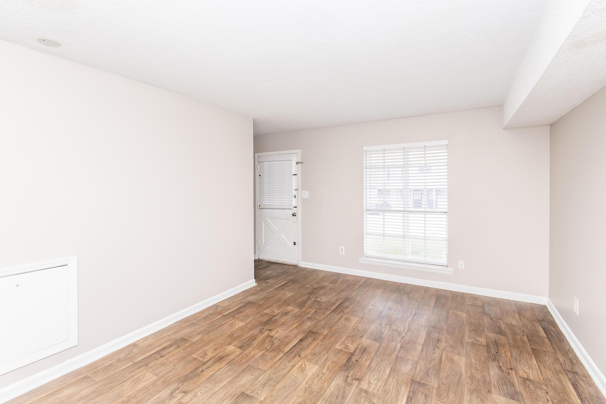 Empty room with light-colored walls and wooden flooring. A door on the left side leads outside, and a window with white blinds lets in natural light. No furniture or decor is present, creating a spacious and minimalist feel.