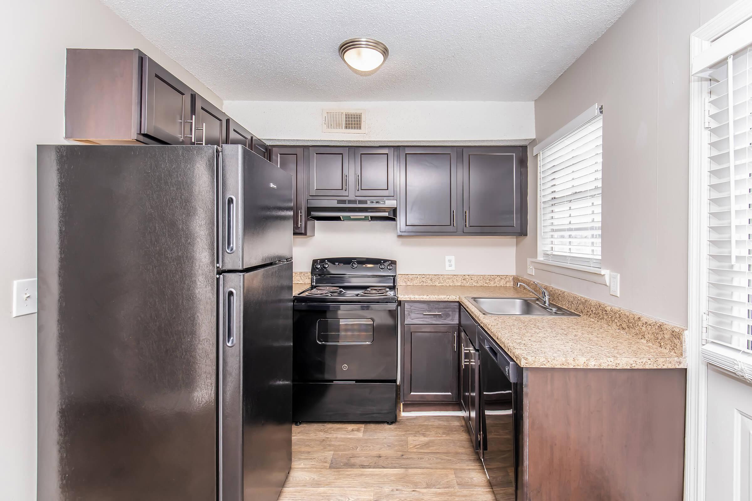 A modern kitchen featuring dark wood cabinets, a black refrigerator, and matching appliances. The countertop is made of light-colored granite, and there is a window providing natural light. The flooring is wood-like, creating a cozy atmosphere.
