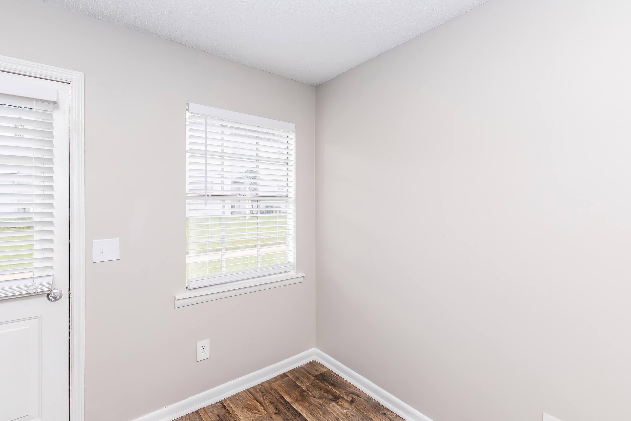 Interior view of a light-colored room with a window featuring white blinds. The floor is wooden, and there is a door on one side. The walls are painted in a soft shade, creating a bright and airy atmosphere. The corner of the room is empty, suggesting potential for decor or furniture.