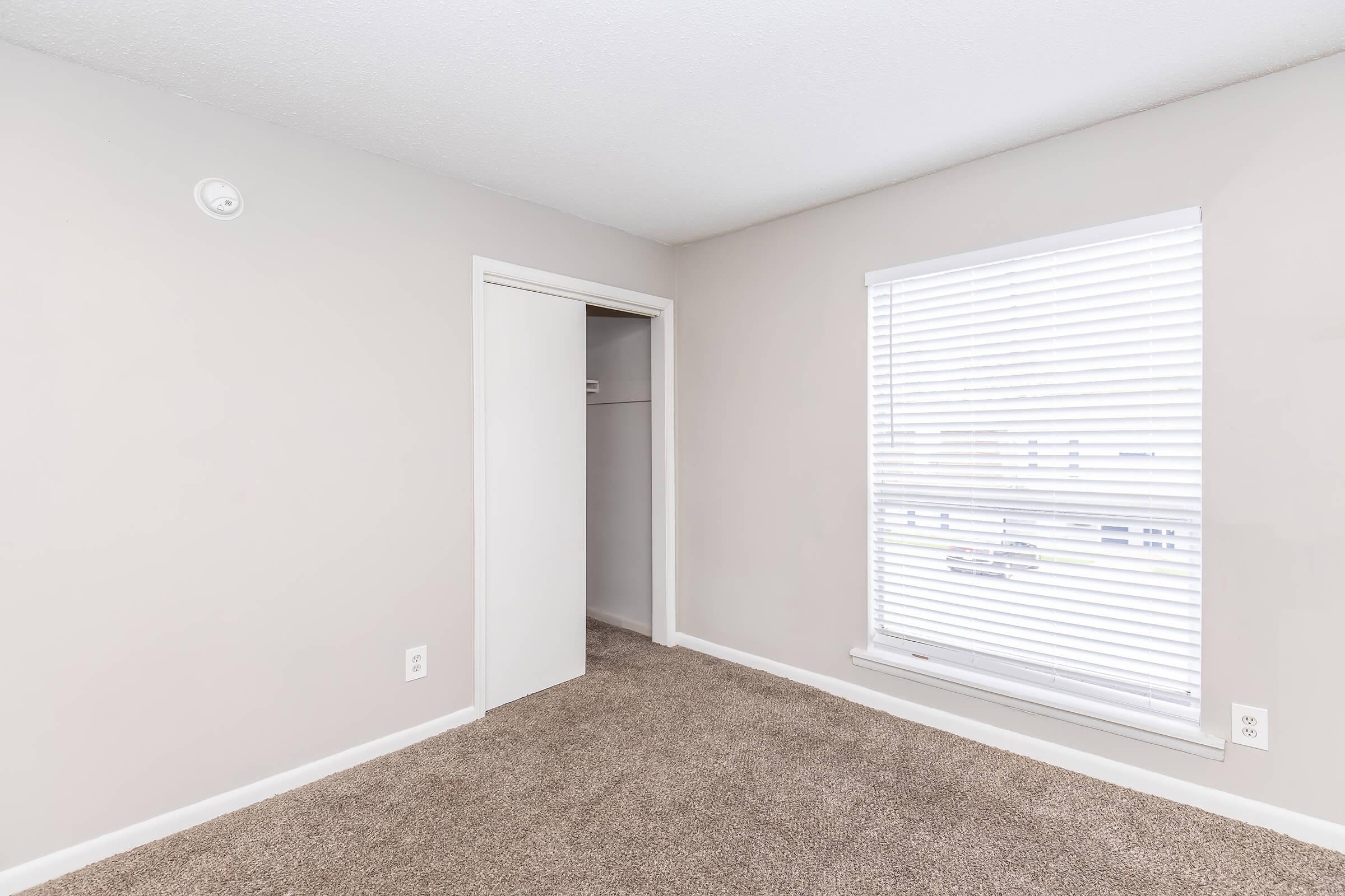 A small, empty bedroom with light beige walls and carpet. The room features a closed door leading to a closet, a window with white blinds allowing natural light, and a simple ceiling fixture. The overall atmosphere is clean and uncluttered, suitable for personal decoration.