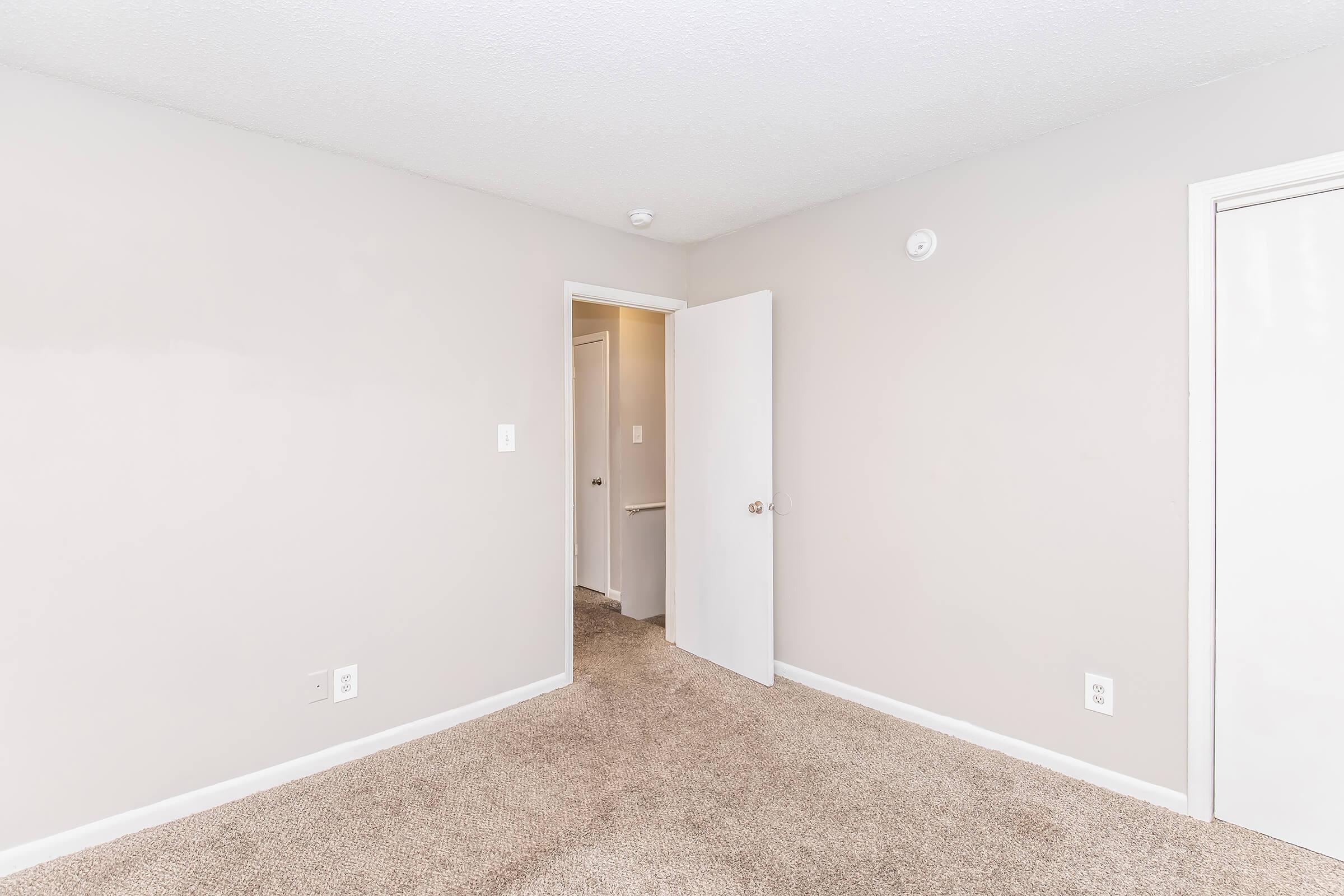 A small, empty room with light beige walls and carpeted flooring. There is a white door partially open leading to a hallway, and another door to the right. The room is well-lit, creating a clean and minimalistic atmosphere. No furniture or decorations are present.