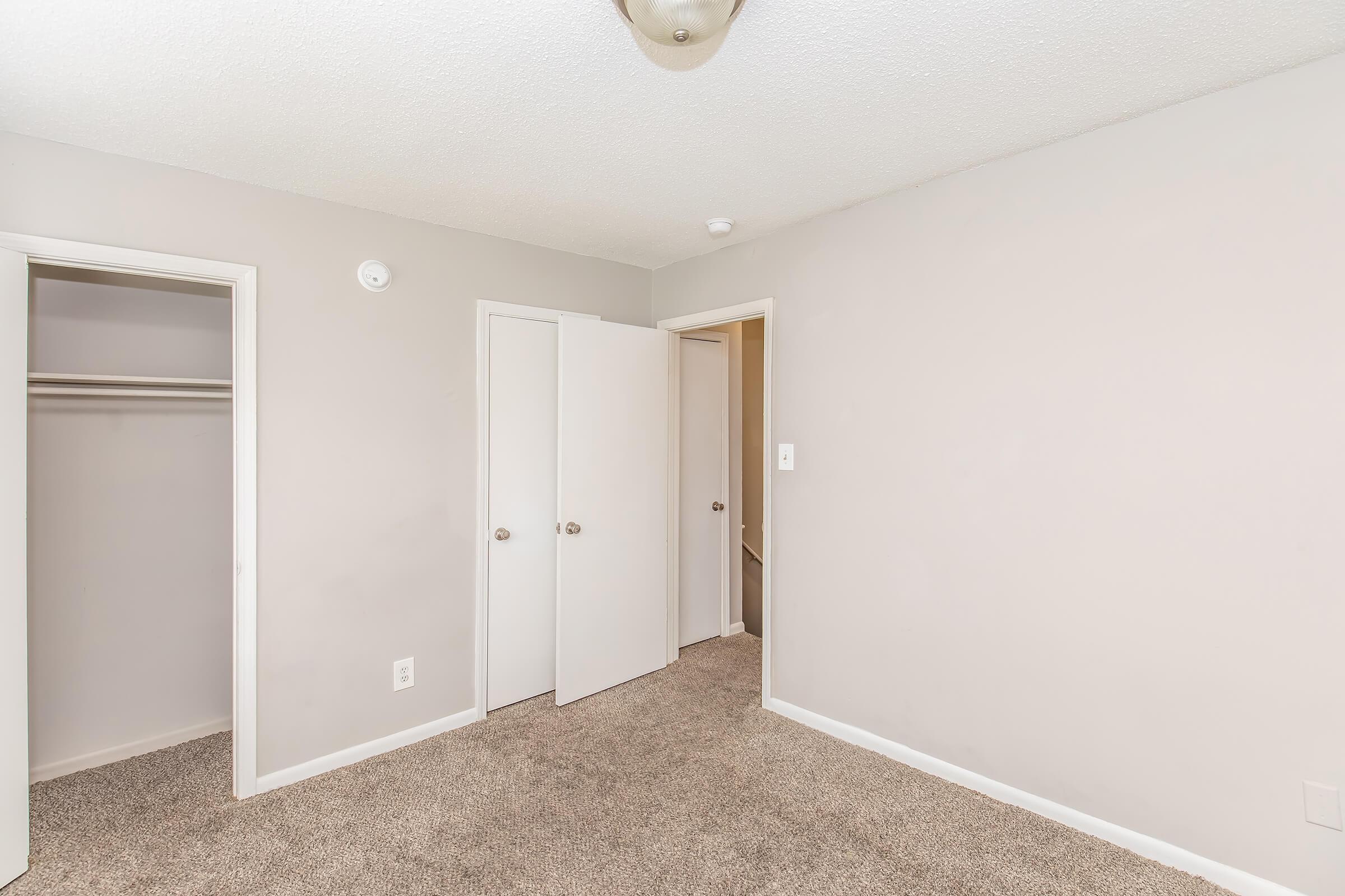 A simple, light-colored bedroom featuring two closed doors leading to closets and a bathroom. The walls are painted in a soft gray tone, and the floor is covered with plush carpeting. A round ceiling light fixture is visible, illuminating the room.