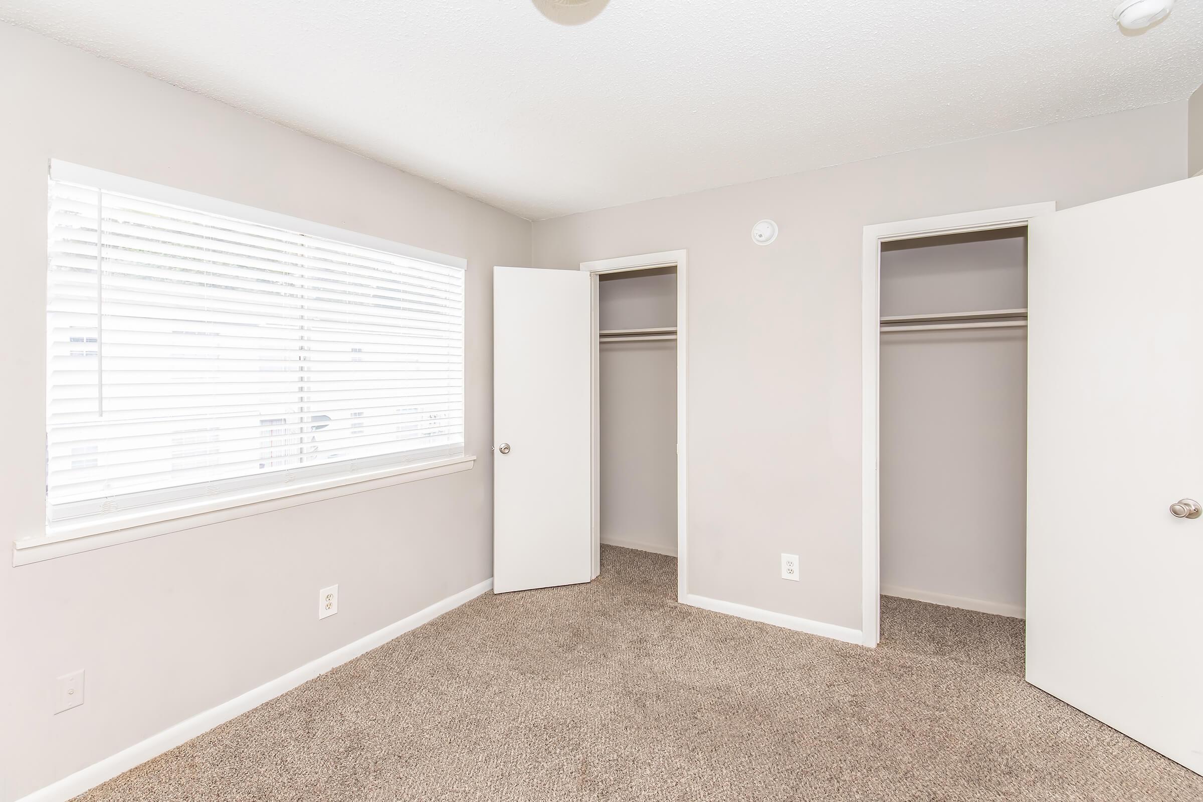 A well-lit, empty bedroom featuring light beige walls and carpeted flooring. Two open closet doors are visible on the right, and a window with white blinds allows natural light to enter the room. The space is minimalistic and ready for personal furnishings.