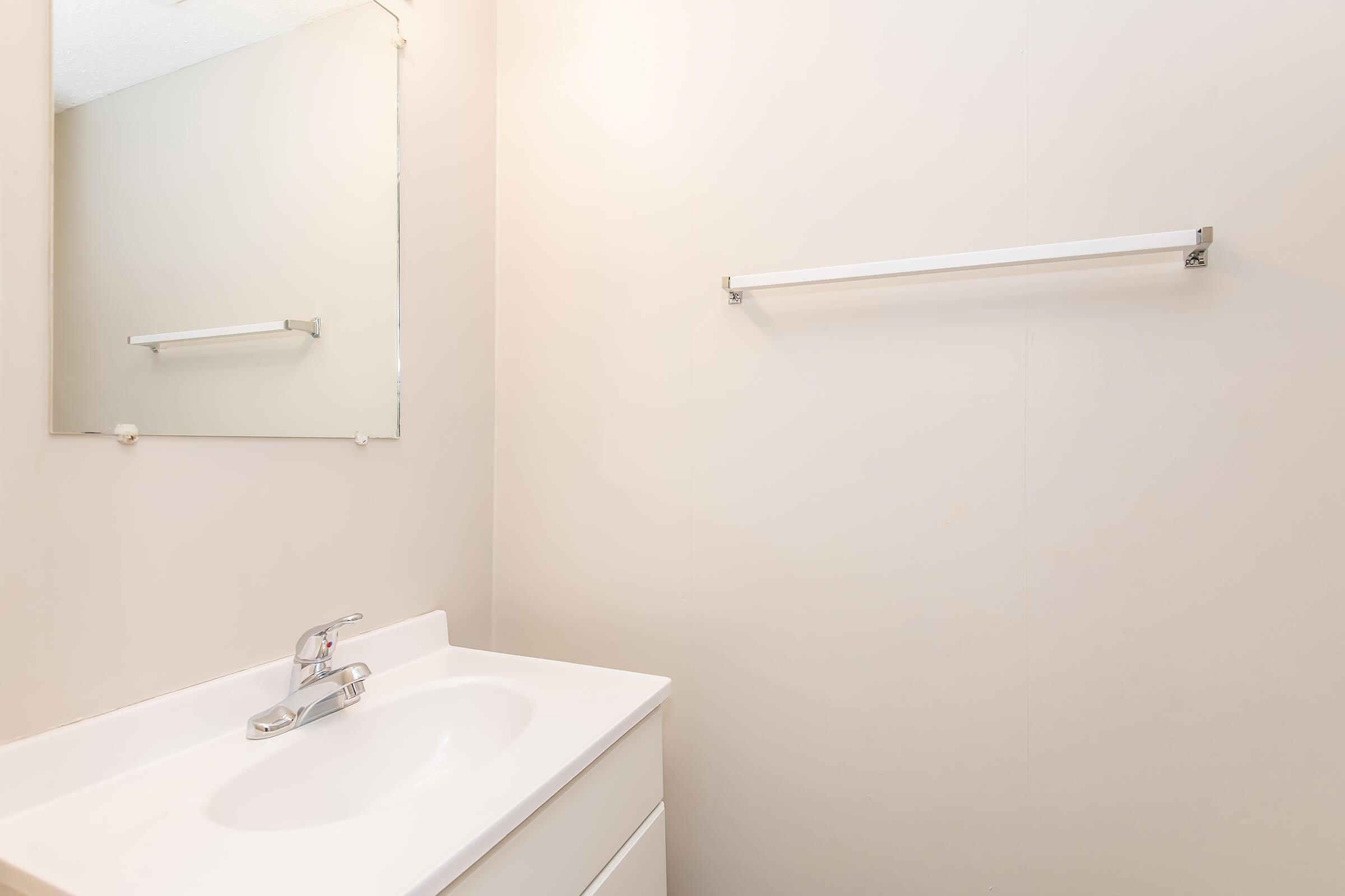 A clean, minimalist bathroom featuring a white sink with a modern faucet. There is a large mirror above the sink and a simple towel bar on the wall. The walls are light-colored, creating a bright and airy atmosphere. The overall design emphasizes simplicity and functionality.