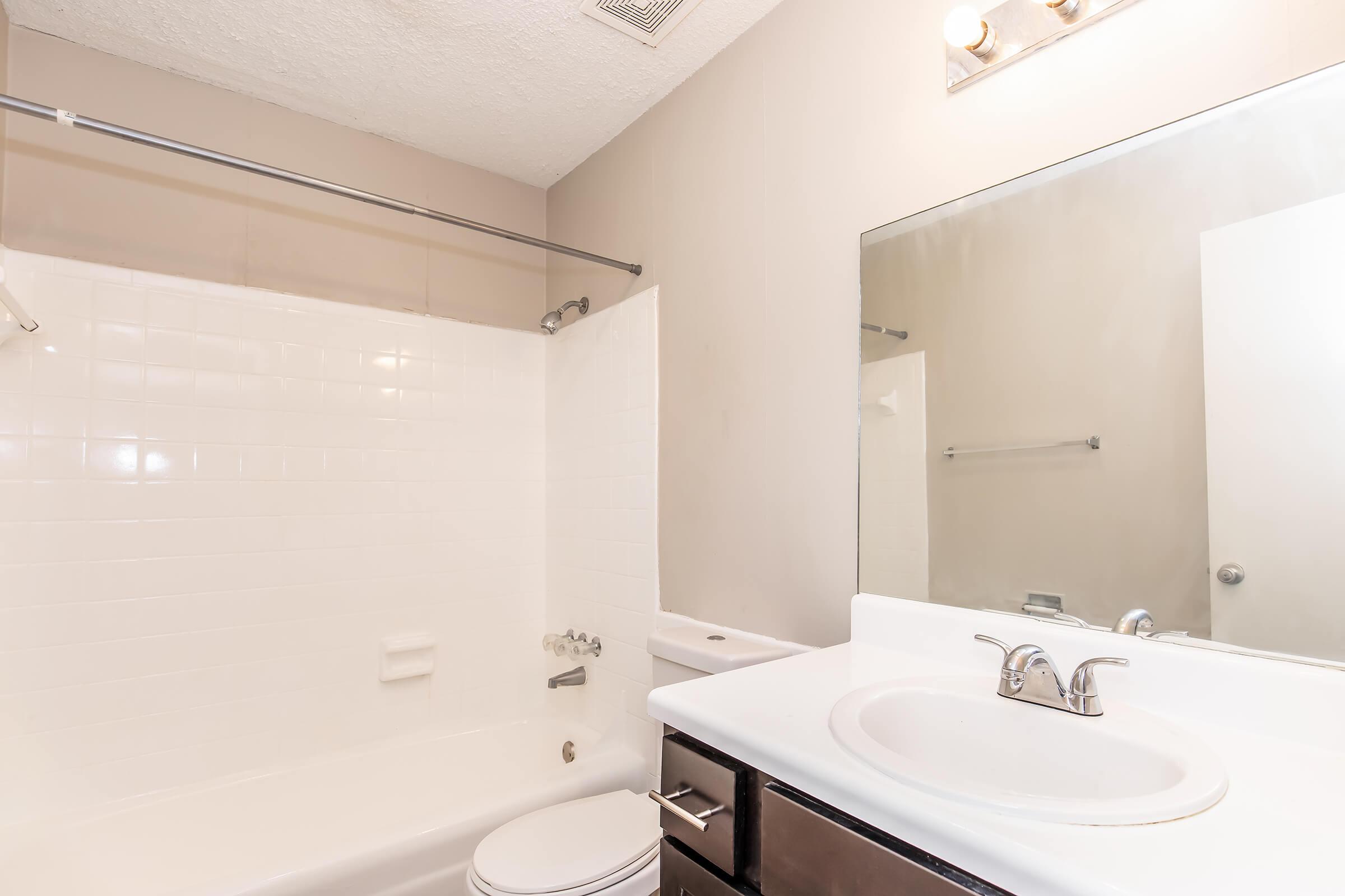 Bright and clean bathroom featuring a white tiled bathtub with a shower, a modern sink with a sleek faucet, and a large mirror above the sink. The walls are a neutral color, and there's a simple rod for towels. The space looks well-maintained with minimal decor.