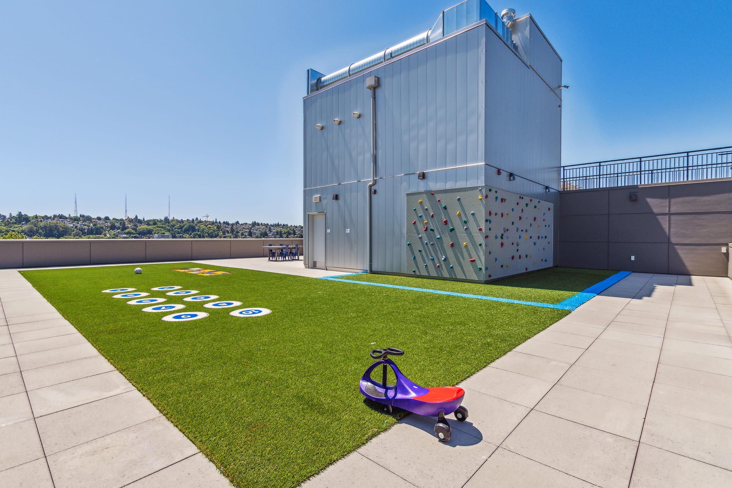 A rooftop play area featuring artificial grass, a climbing wall, and large colorful letters on the ground. A small purple and red ride-on toy car is parked on the grass. The background shows a clear blue sky and a cityscape.