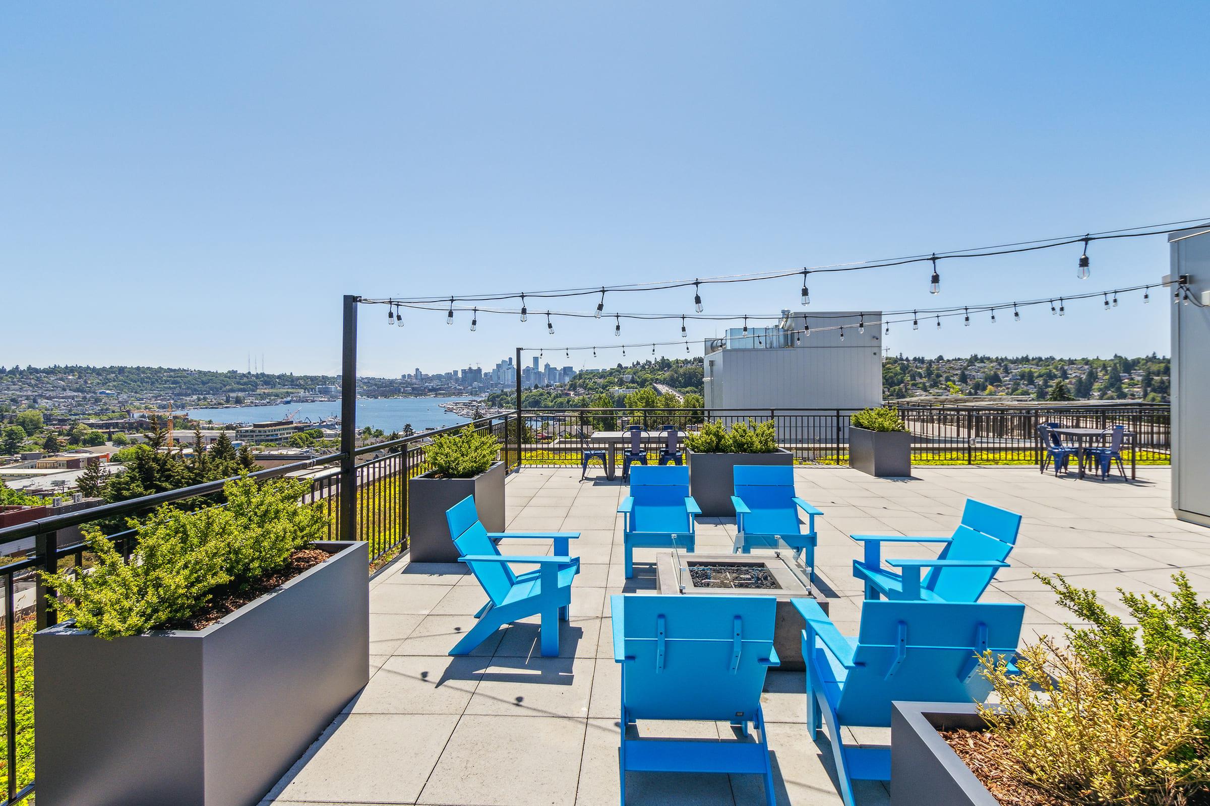 A rooftop terrace featuring blue seating and planters, overlooking a scenic view of a city skyline and water. String lights are hung above, creating a welcoming atmosphere for gatherings. The space is bright and sunny, with greenery and modern furnishings enhancing the outdoor setting.