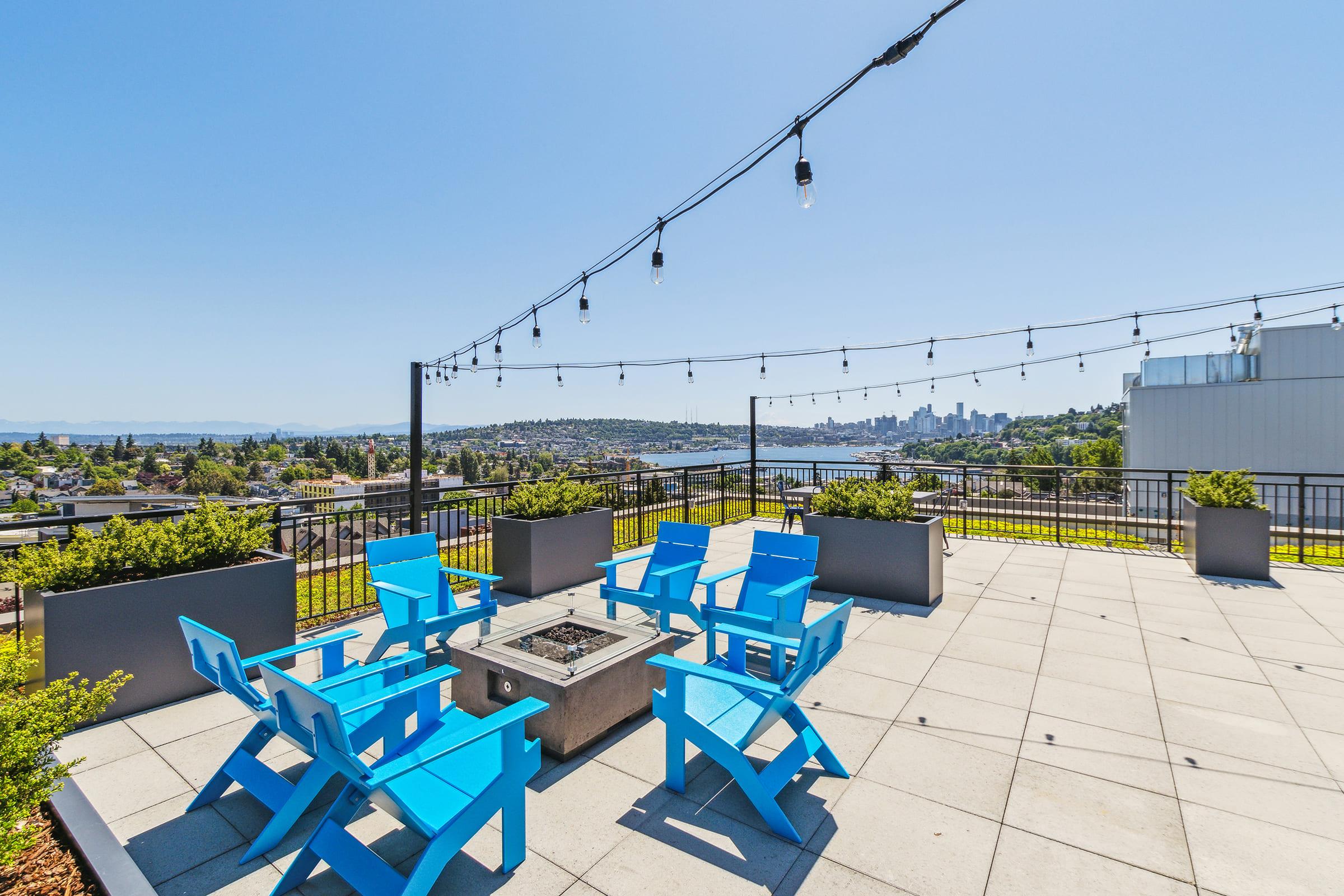 A rooftop terrace featuring bright blue Adirondack chairs arranged around a fire pit. The scene includes string lights overhead and planters, with a scenic view of a city skyline and mountains in the background under a clear blue sky.