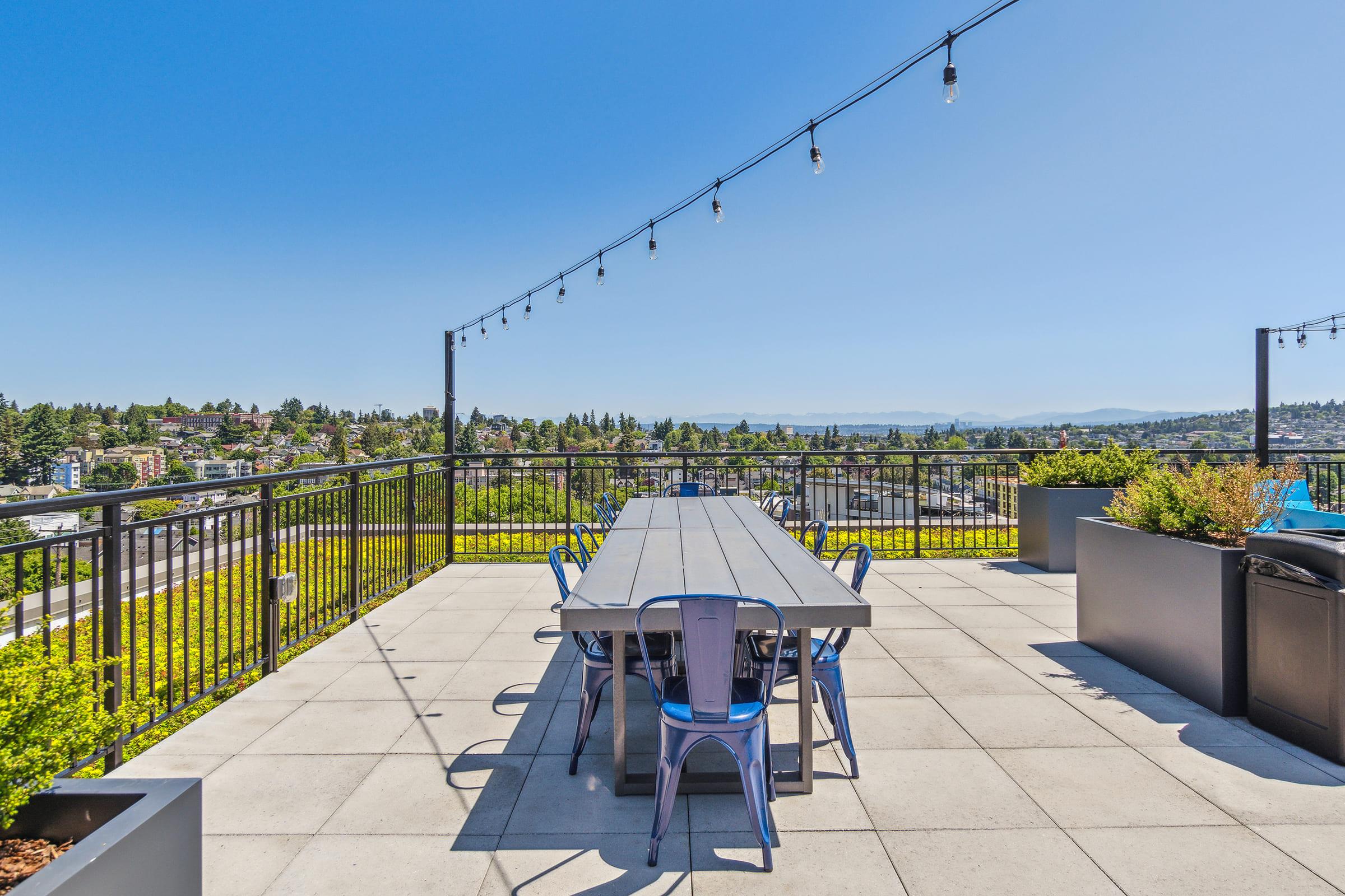 A rooftop terrace featuring a long, gray dining table surrounded by metal chairs, with string lights overhead. Lush greenery and contemporary planters frame the edges, and a panoramic view of the city stretches out in the background under a clear blue sky.