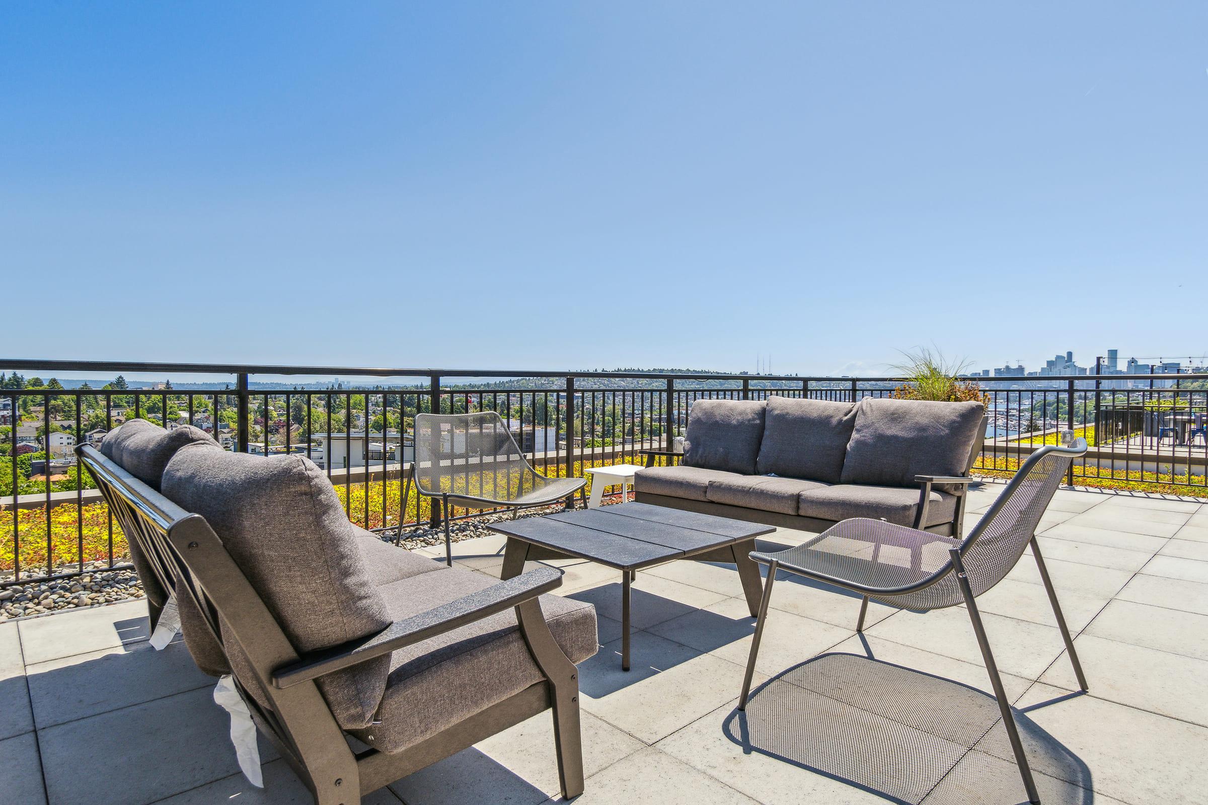 A modern outdoor patio featuring a gray sectional sofa, a glass coffee table, and two lounge chairs. The space is surrounded by a black railing with a panoramic view of the city skyline and lush greenery under a clear blue sky. Ideal for relaxation and entertaining.