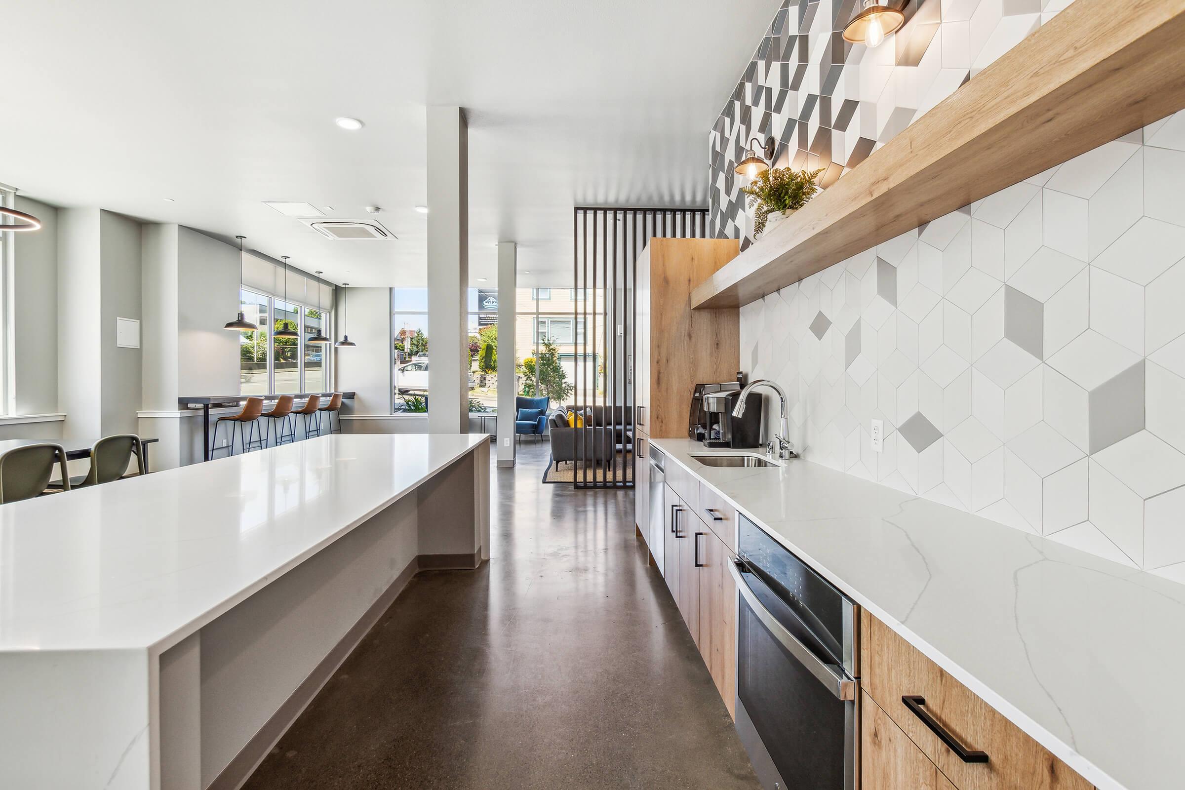 Modern kitchen interior featuring a long white countertop, wooden cabinetry, and geometric tile backsplash. The space includes an electric stove, coffee maker, and bar seating in the background, with large windows allowing natural light to fill the room. The floor is concrete, adding to the contemporary aesthetic.