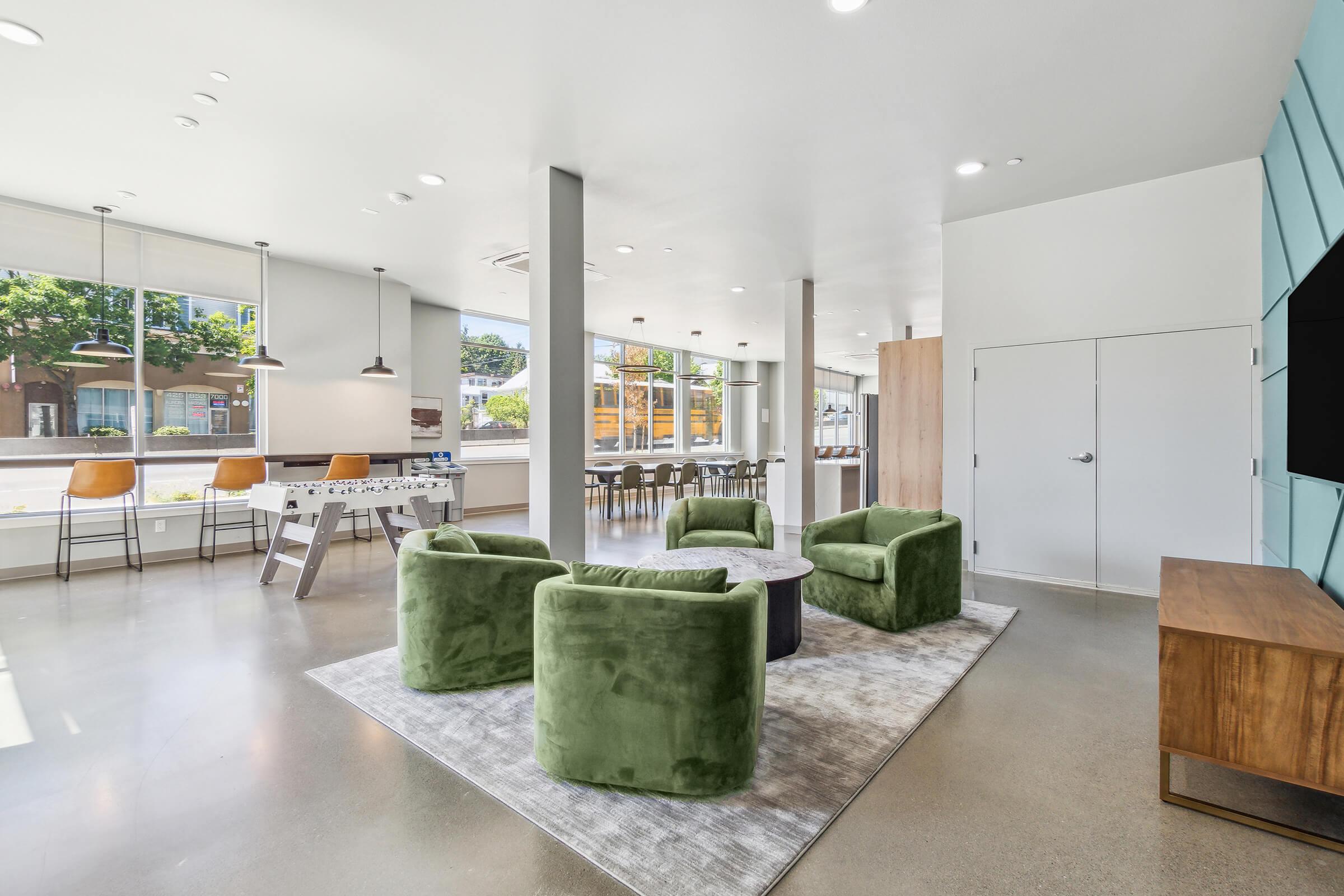 A modern, bright lounge area featuring four green upholstered chairs arranged around a circular coffee table. Large windows provide natural light and a view of the outside. In the background, there are bar stools at a high table and a wooden sideboard against a wall. The floor is polished concrete.