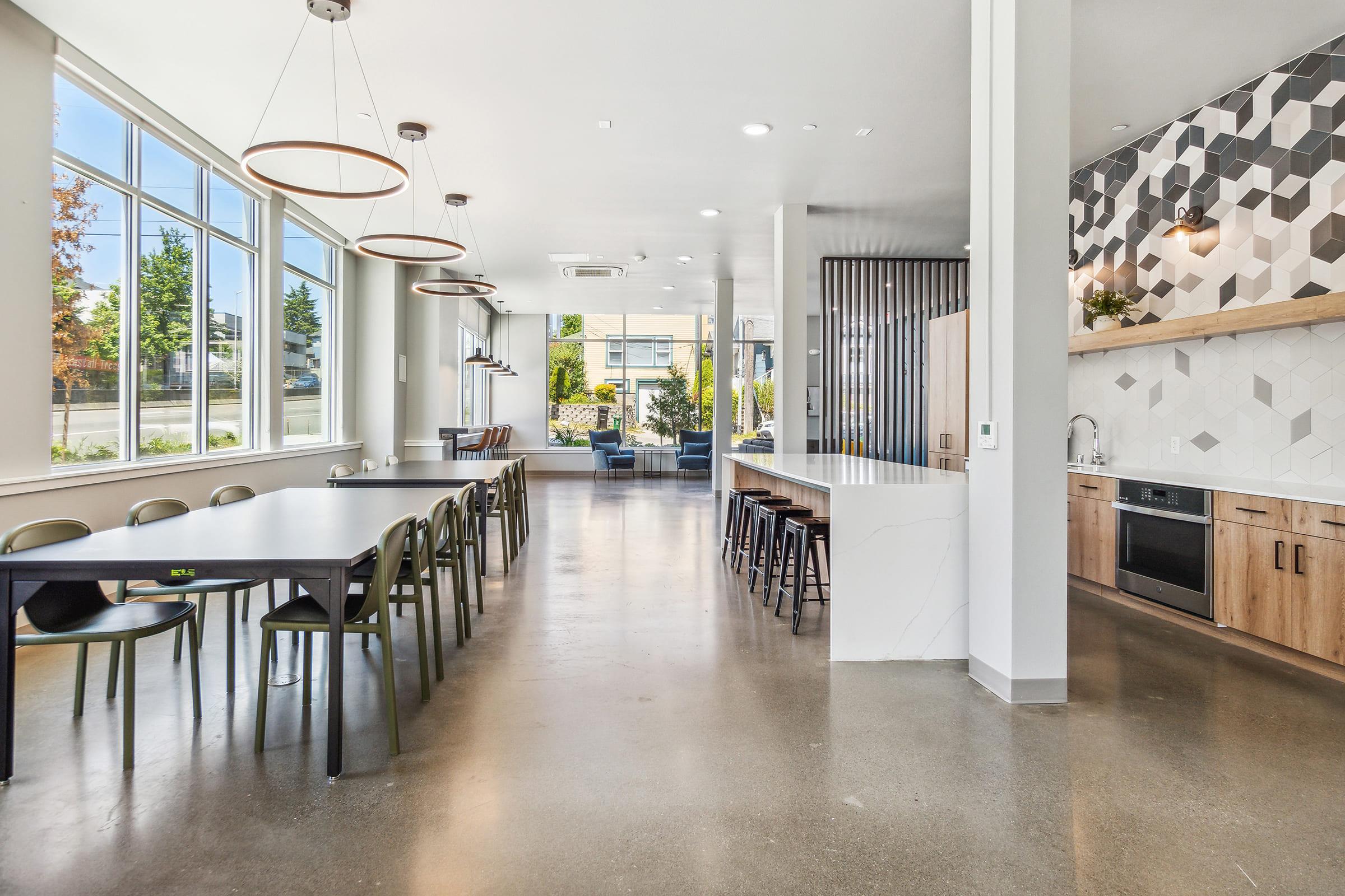 A modern communal space featuring a large dining table and individual seating. The room has ample natural light from large windows, stylish pendant lighting, and a kitchenette with wooden cabinets and a geometric tile backsplash. The polished concrete floor adds a contemporary touch to the design.