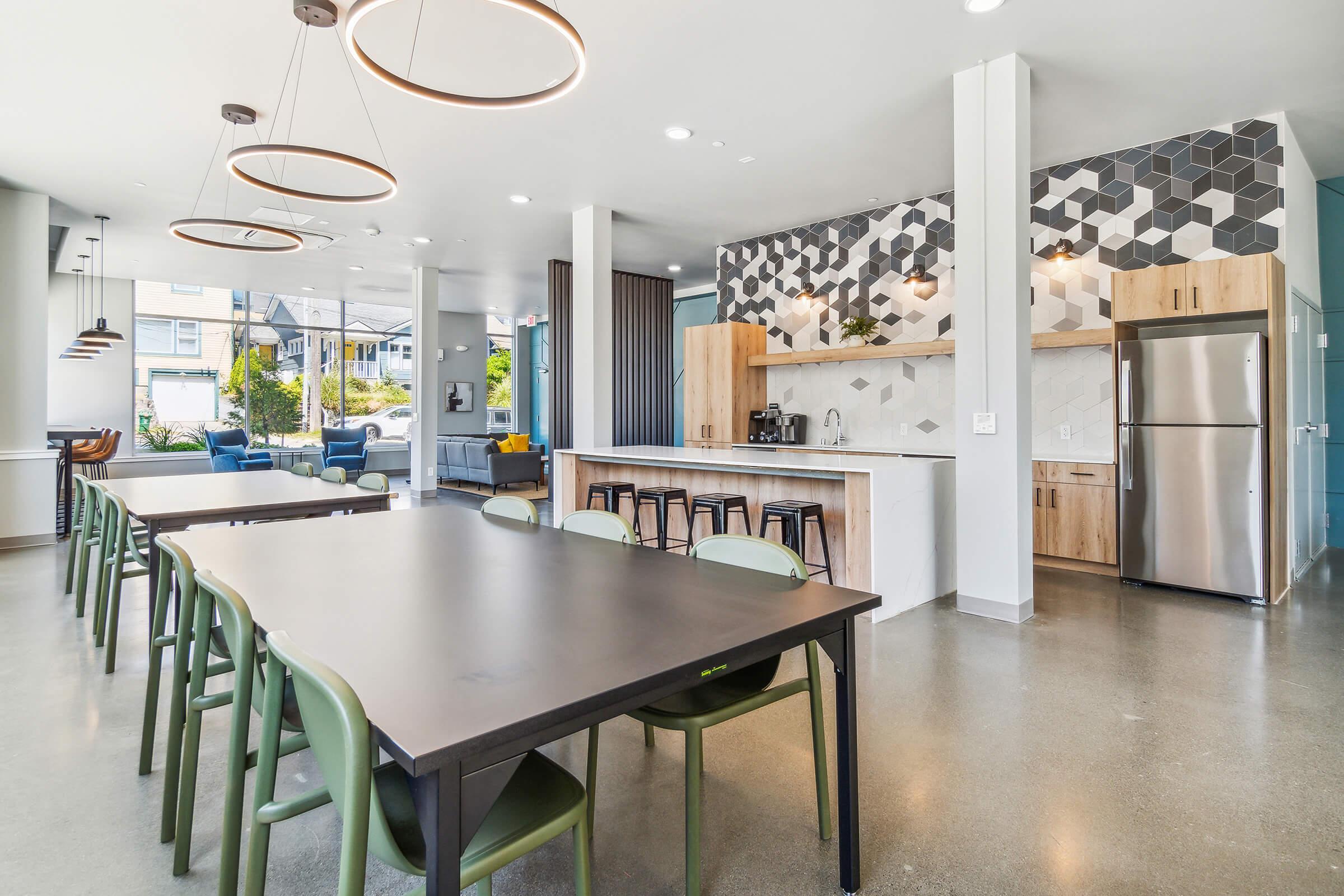 Modern kitchen and dining area featuring a long black table with green chairs, pendant lighting, and hexagonal patterned wall tiles. A kitchenette with wooden cabinetry and stainless steel appliances is visible in the background, alongside large windows that provide natural light.