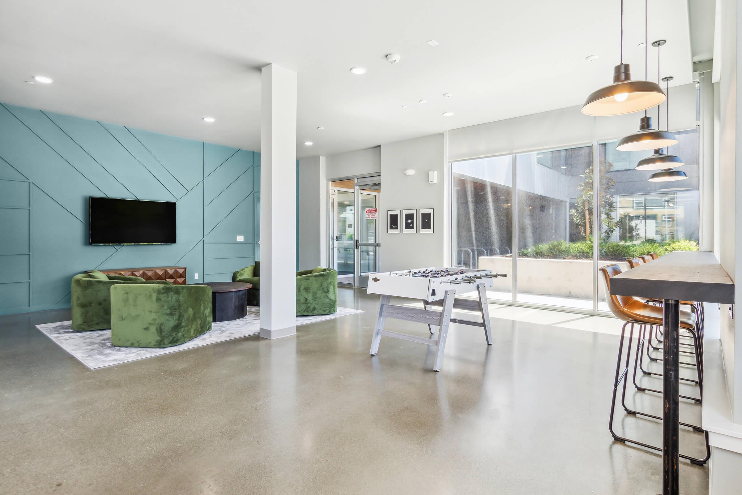 A modern common area featuring green upholstered seating arranged around a rug, a wall-mounted TV, and a foosball table. Large windows let in natural light, and there's a bar-height table with high stools. The decor includes a light blue accent wall and concrete flooring, creating a stylish and inviting atmosphere.