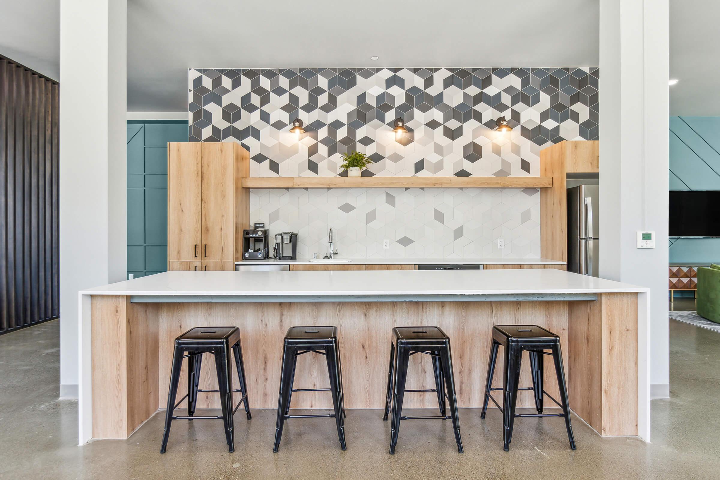 A modern kitchen featuring a geometric tiled backsplash in shades of gray and white. The countertop is white, complemented by wooden cabinetry. Four black metal stools are positioned in front of the counter. Bright lighting fixtures hang above, adding a contemporary feel to the space.