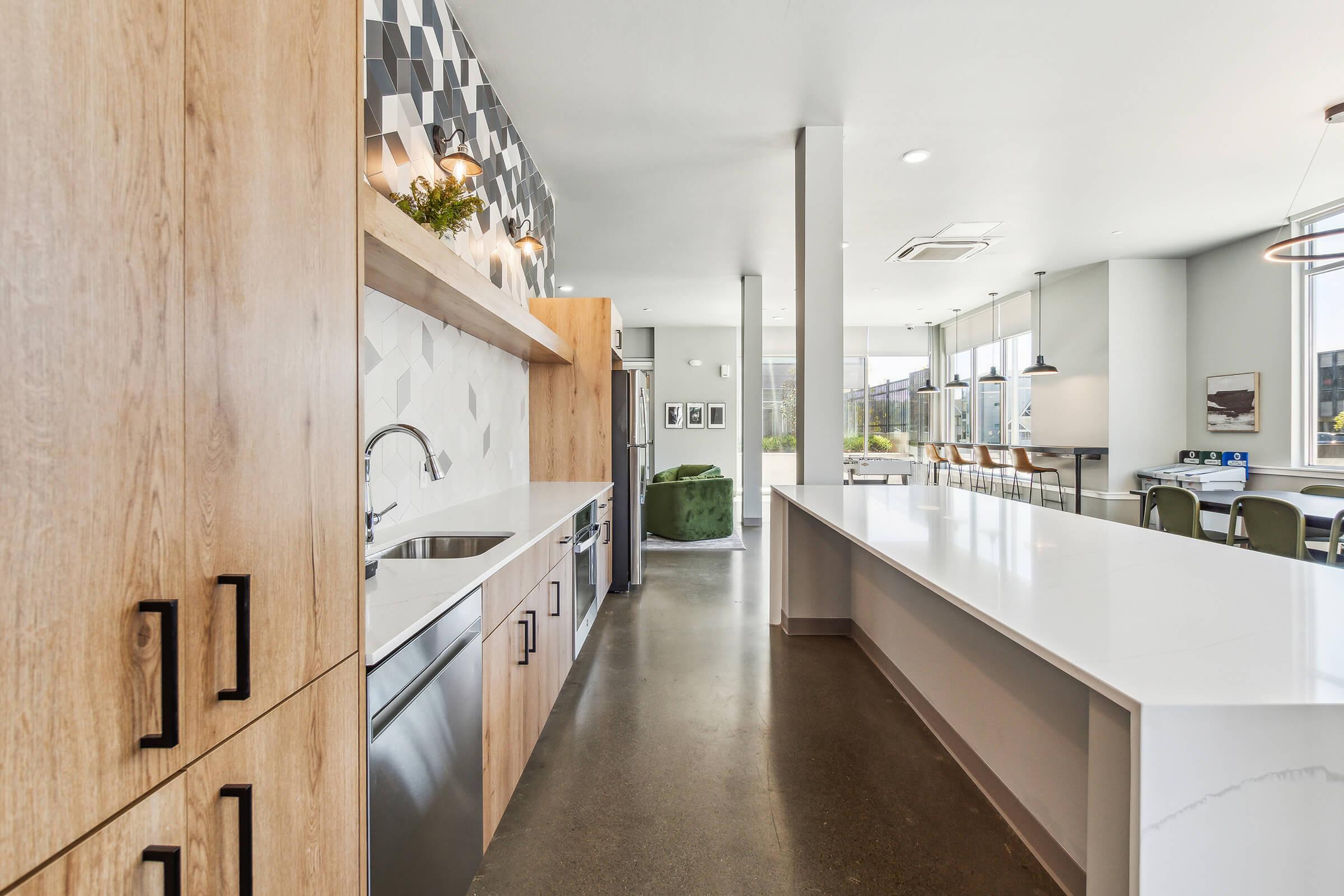 Modern kitchen interior featuring wooden cabinetry, a stainless steel sink, and a sleek countertop. An open layout connects to a dining area with industrial-style lighting and large windows. Neutral colors and geometric wall decor create a contemporary aesthetic.