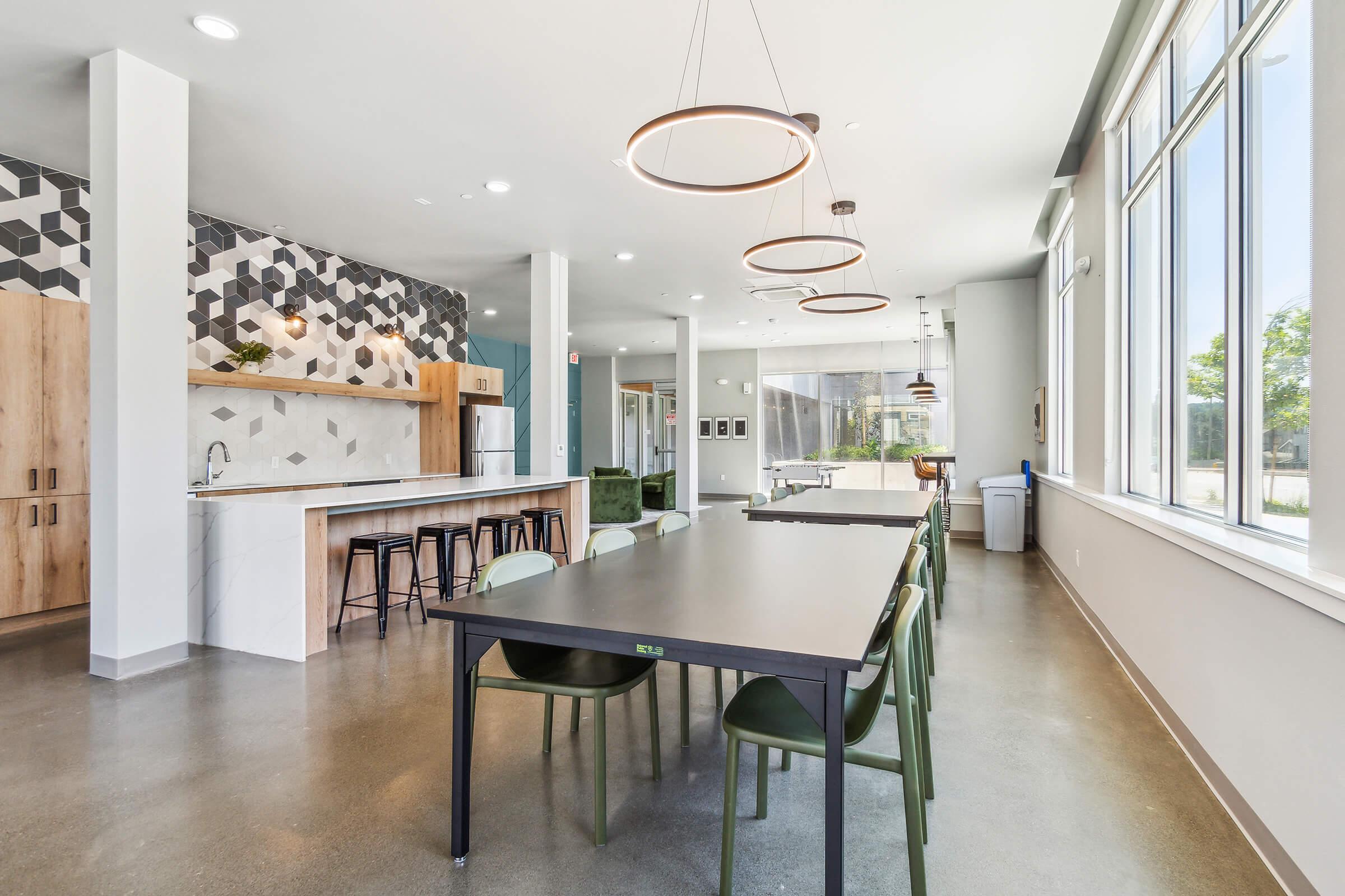 A modern kitchen and dining area featuring a long black table with green chairs, pendant lighting, and a patterned backsplash. Large windows provide natural light, and the space includes wooden cabinets, a stainless steel refrigerator, and a small seating area with greenery. The floors are polished concrete.