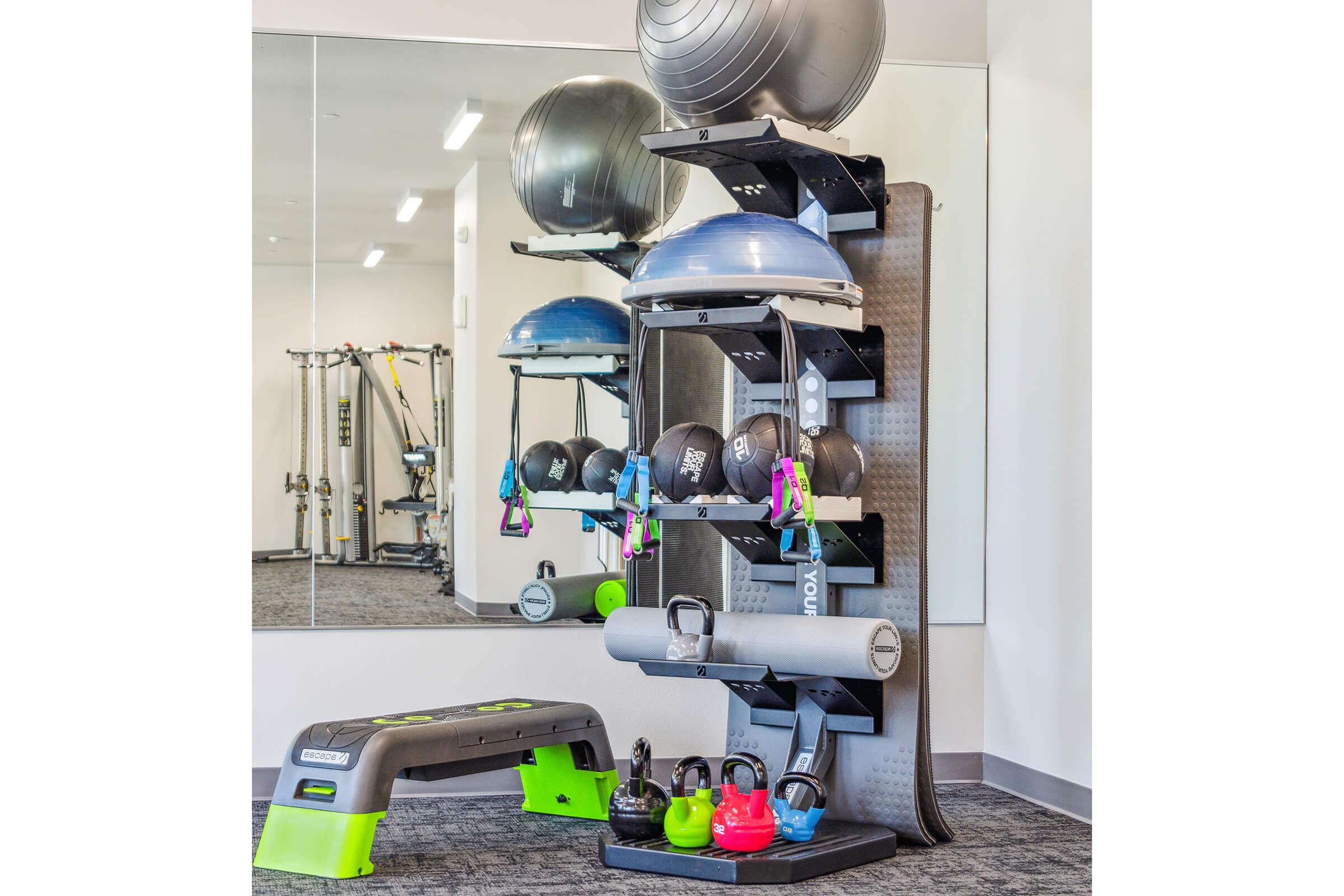 A fitness equipment display featuring a rack with stability balls, medicine balls, resistance bands, and kettlebells. A grey step platform is positioned in front, and a mirror reflects the setup, creating a spacious workout environment with a clean, modern aesthetic.