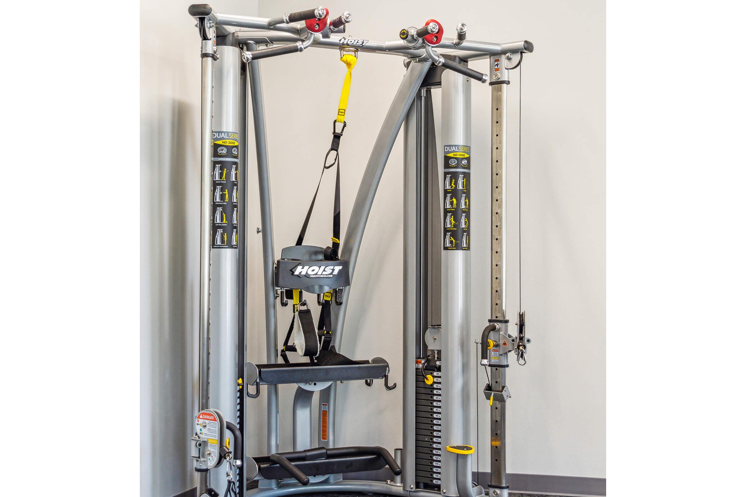 A multi-functional exercise machine featuring various pulley systems, adjustable weight settings, and a central bench with a harness. The machine is designed for strength training and includes multiple attachment options for a range of exercises.