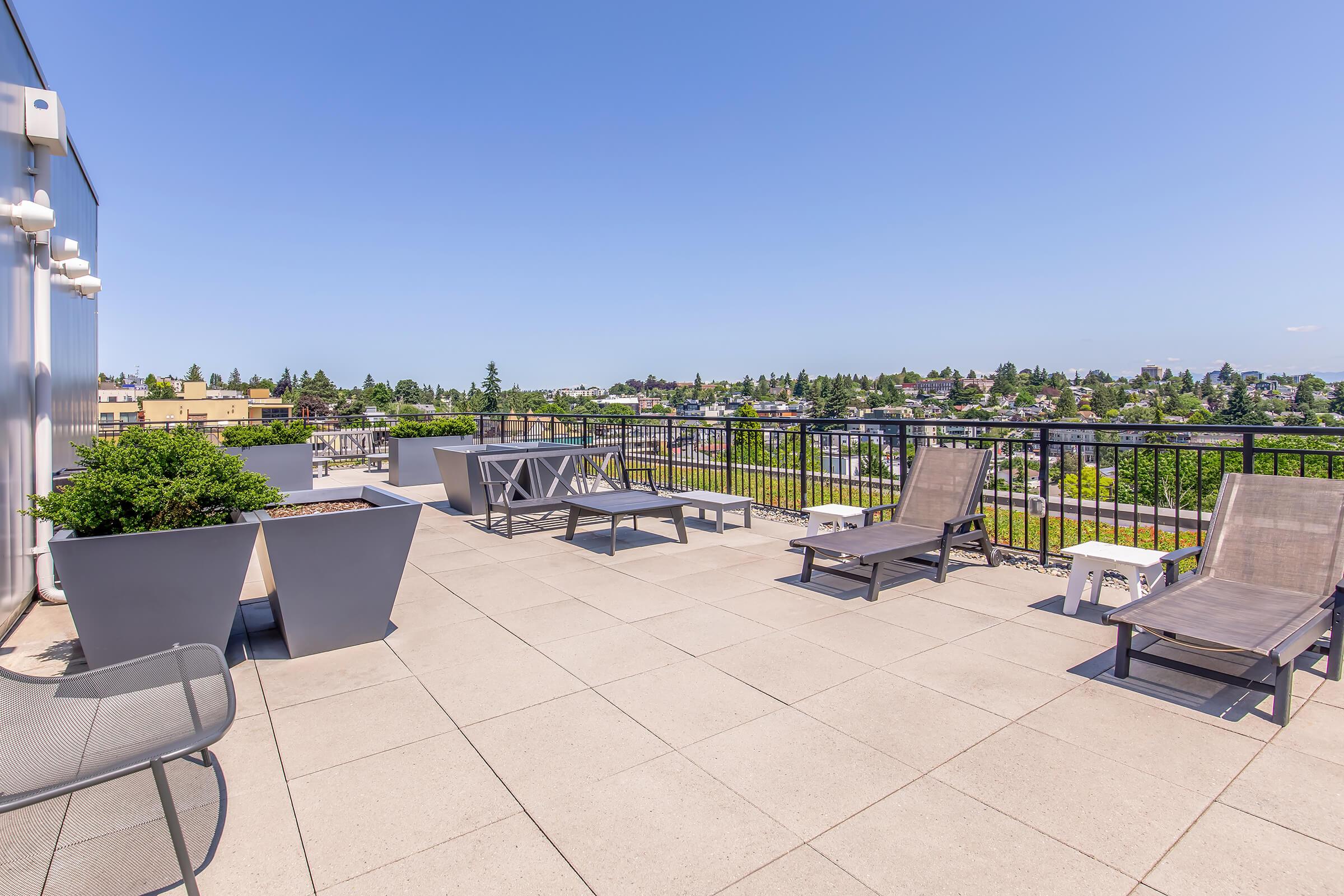 A rooftop terrace with modern lounge chairs and planters, featuring a wide view of the surrounding urban landscape under a clear blue sky. The space is designed for relaxation and offers a tranquil environment amidst the city.