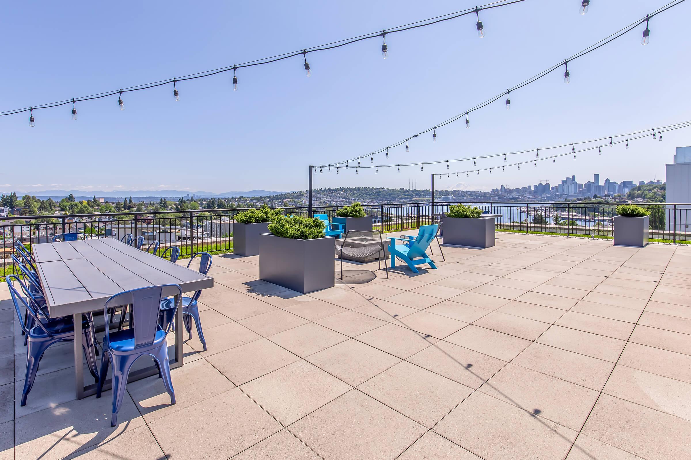 A spacious rooftop terrace with a large dining table, blue chairs, and cozy seating areas. The space is adorned with string lights and overlooks a scenic view of a city skyline and water in the background, creating a relaxing outdoor atmosphere.