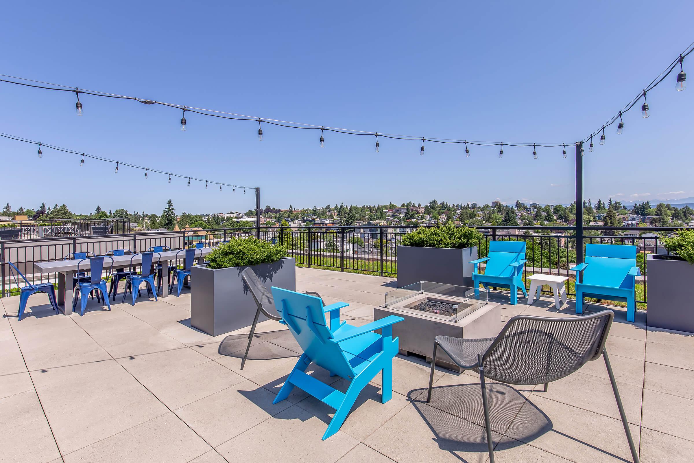 A rooftop space featuring colorful blue lounge chairs, a fire pit, and a dining area with tables and chairs. String lights hang overhead, and the area offers a panoramic view of the surrounding cityscape and greenery under a clear blue sky.