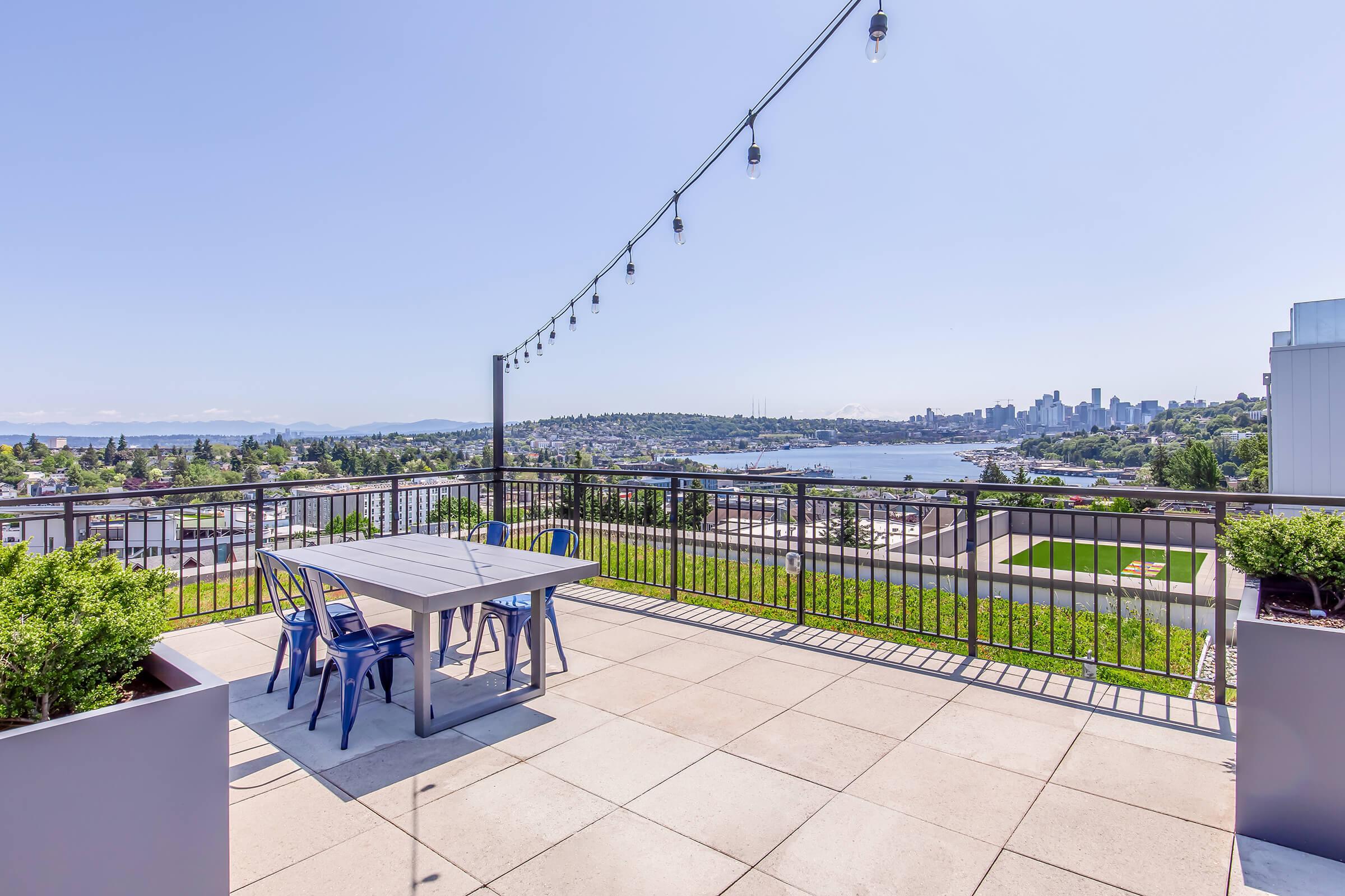 A spacious rooftop terrace with a modern dining table and chairs, overlooking a scenic view of a waterfront cityscape, lush greenery, and string lights illuminating the area on a sunny day.