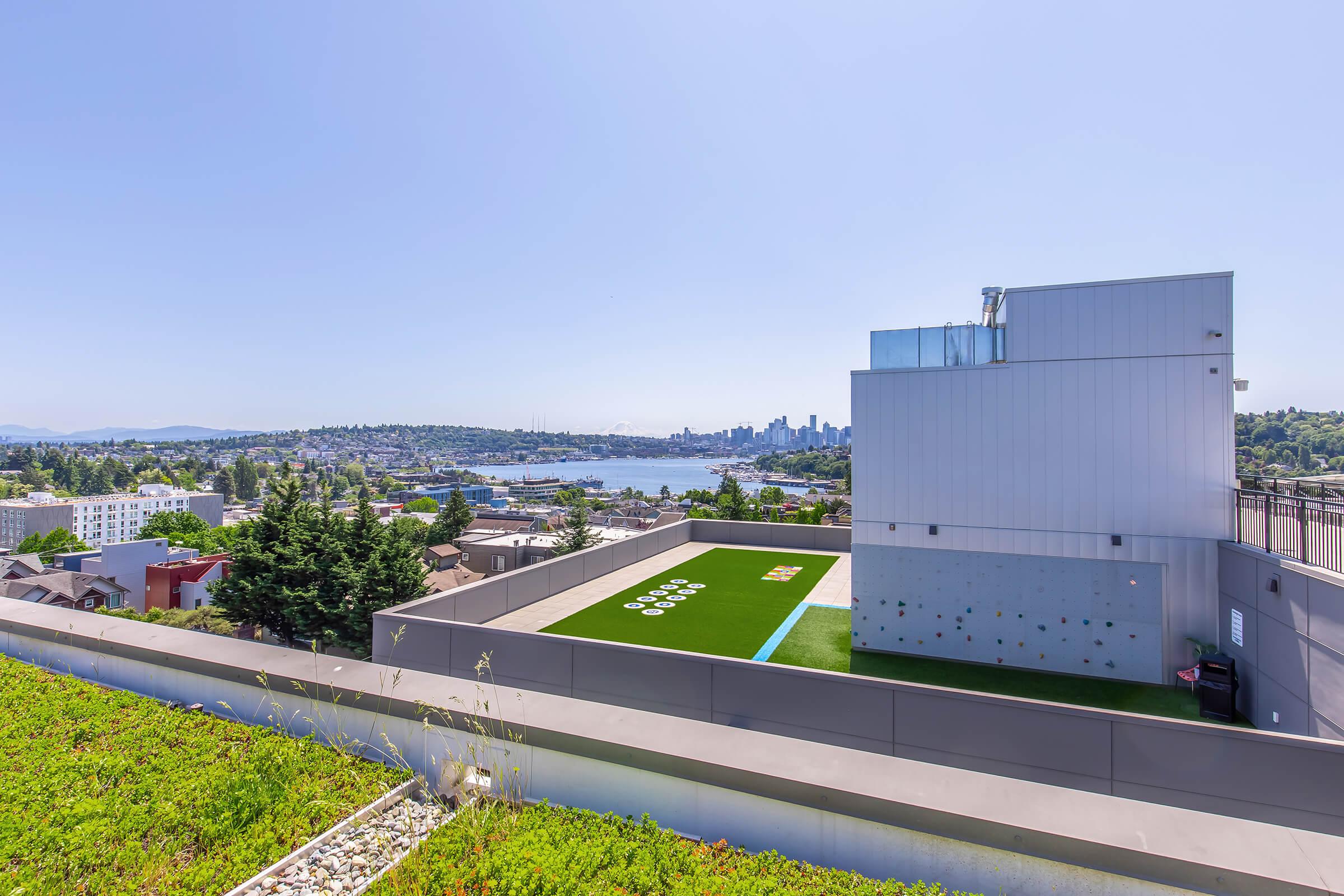 A rooftop view featuring a green space and climbing wall, overlooking a cityscape with buildings and a body of water in the background. Bright blue sky enhances the vibrant scenery, showcasing a mix of urban life and nature.