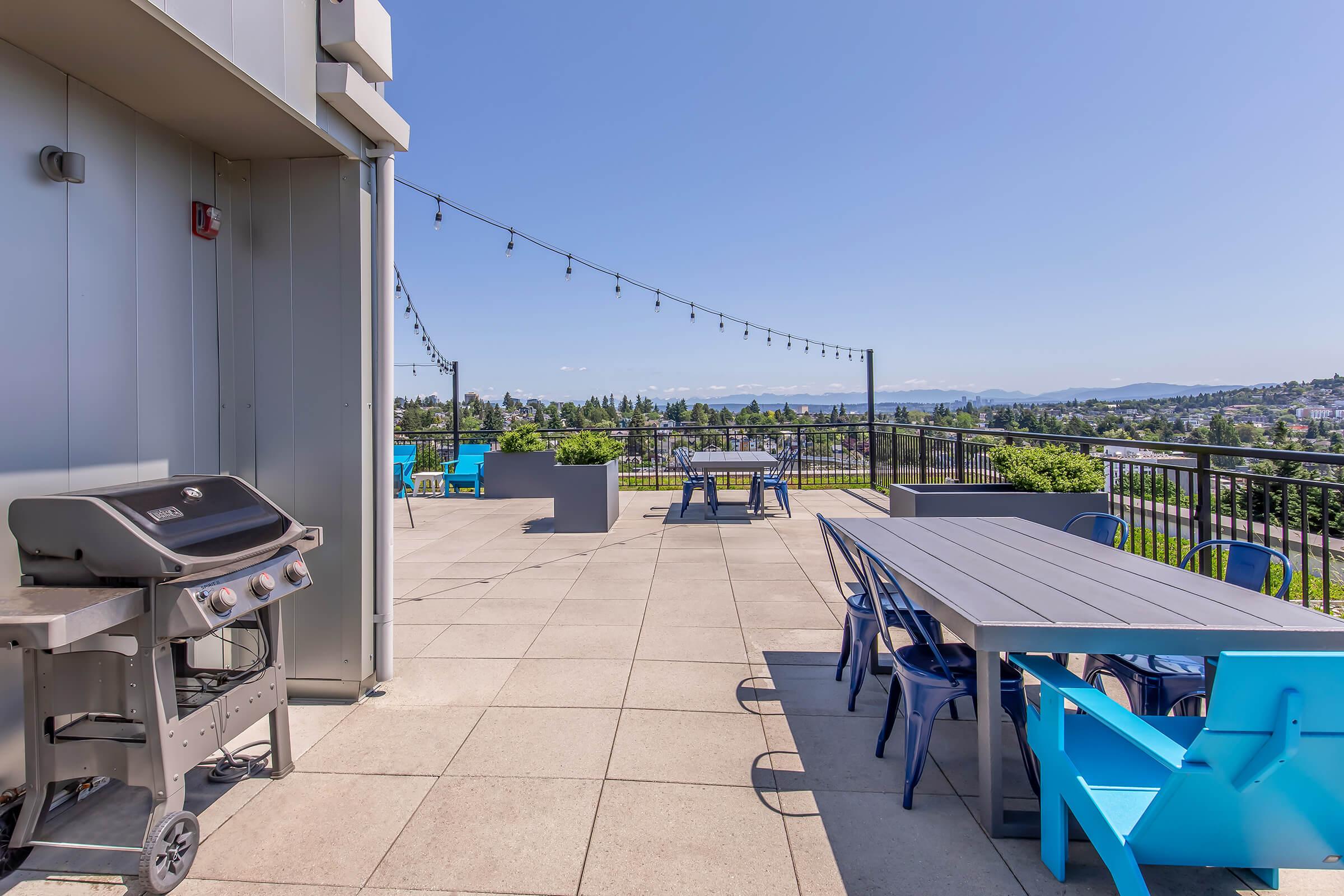 A rooftop patio featuring a stainless steel barbecue grill, a large dining table with blue chairs, and several smaller seating areas. The space is surrounded by a railing and offers a scenic view of the landscape and sky, with string lights above for ambiance.
