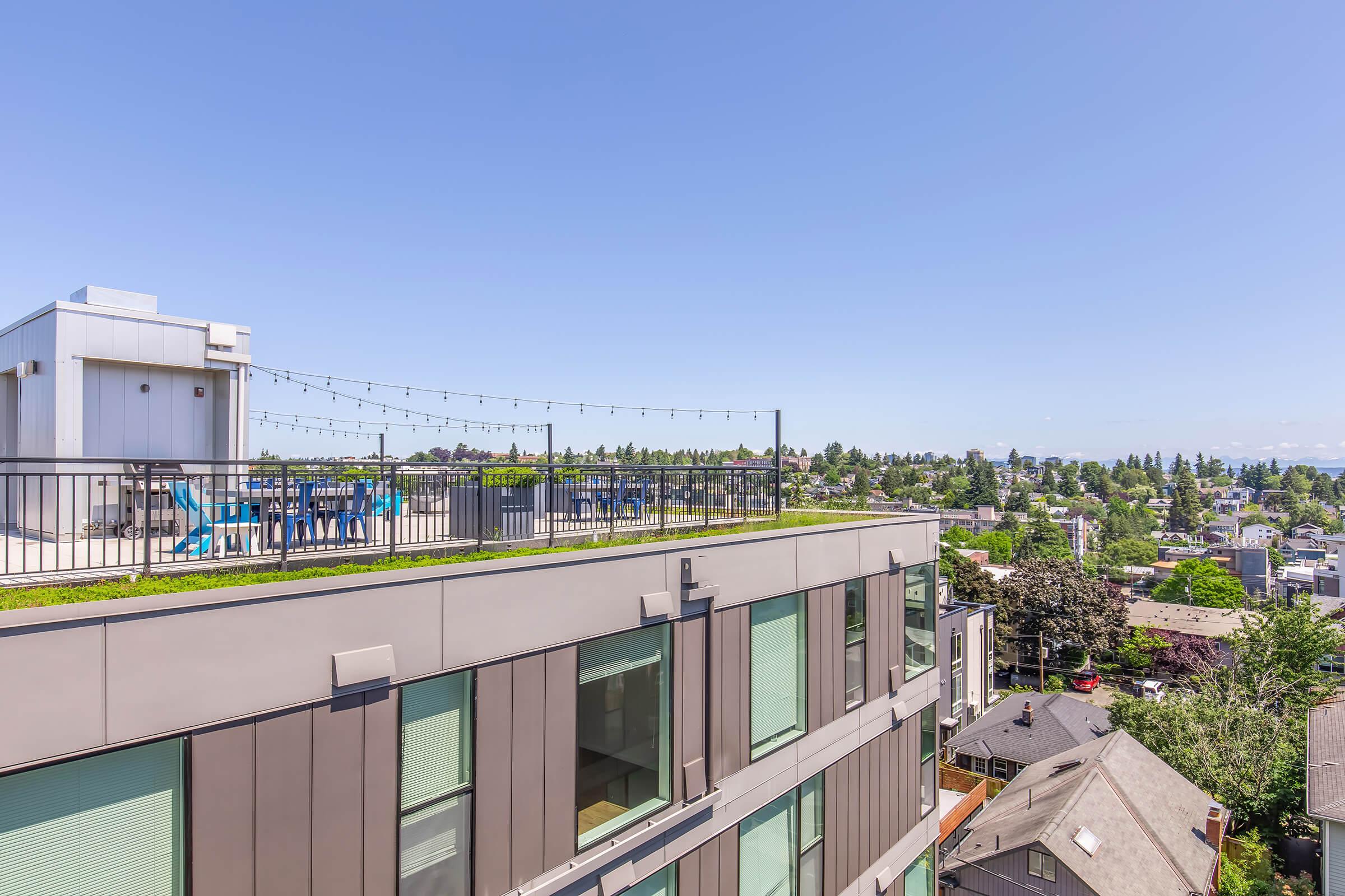 A view from a rooftop balcony featuring a green rooftop patio with blue chairs and string lights. The background includes a cityscape with various buildings, trees, and a clear blue sky.