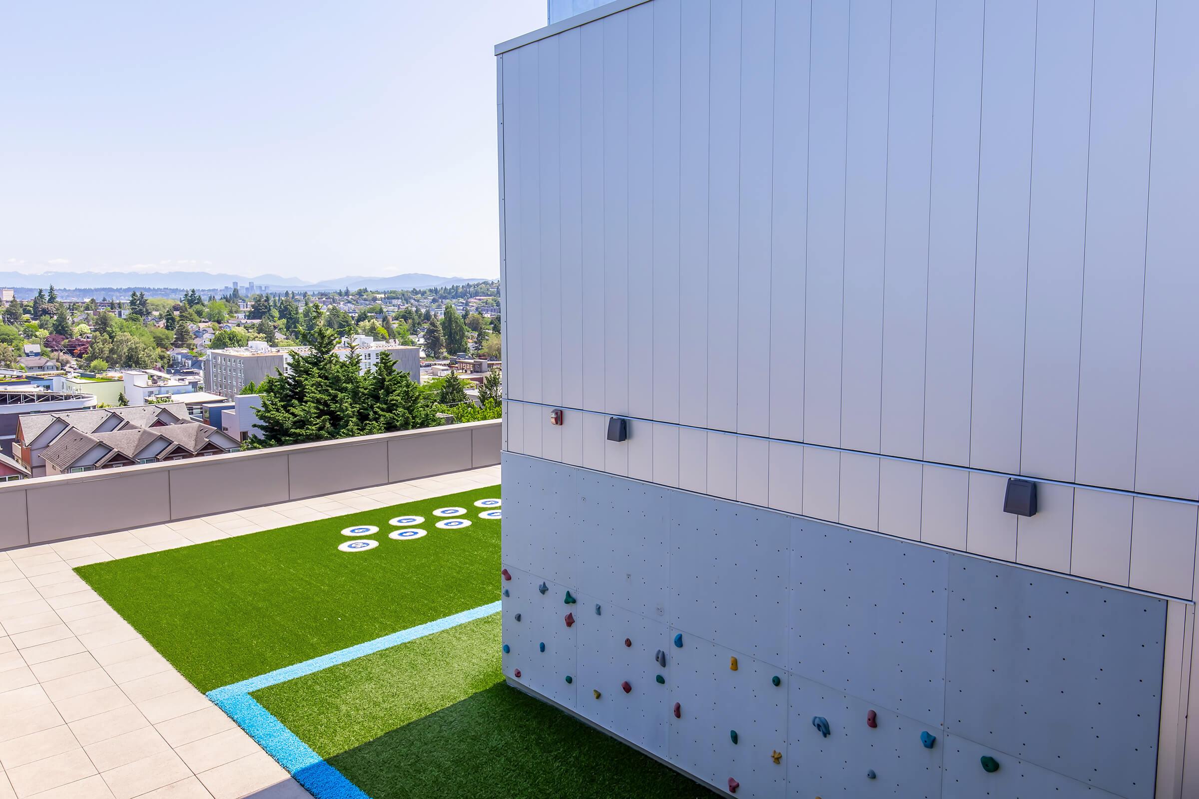 Rooftop area featuring a climbing wall and artificial grass. The scene overlooks a cityscape with trees and distant mountains in the background under a clear blue sky. Circular white pads are scattered on the grassy area, providing a leisure space.