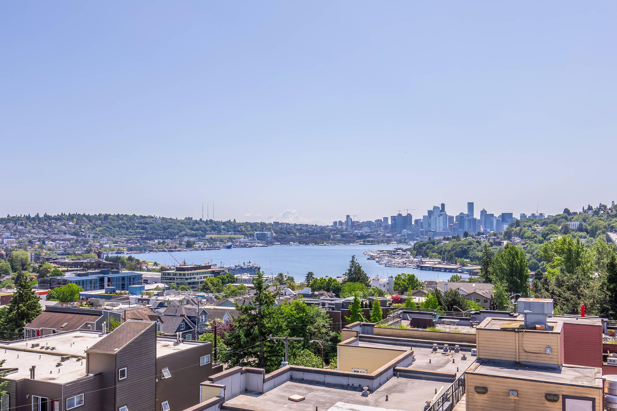 A panoramic view of a vibrant cityscape, featuring a body of water surrounded by green trees and residential buildings. In the distance, modern skyscrapers of a downtown area are visible against a clear blue sky, with mountains peeking through in the background. The scene is bright and sunny, showcasing a lively urban environment.
