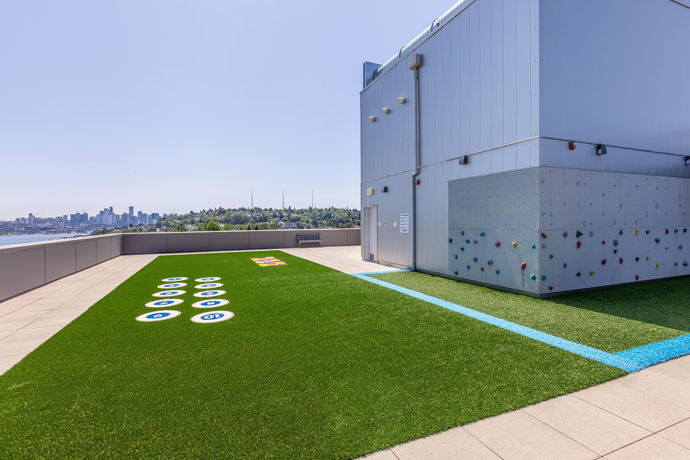 A rooftop area featuring synthetic grass with various colorful circular markings on the ground and a climbing wall on the side. The background displays a city skyline under a clear blue sky, providing a vibrant and recreational atmosphere.