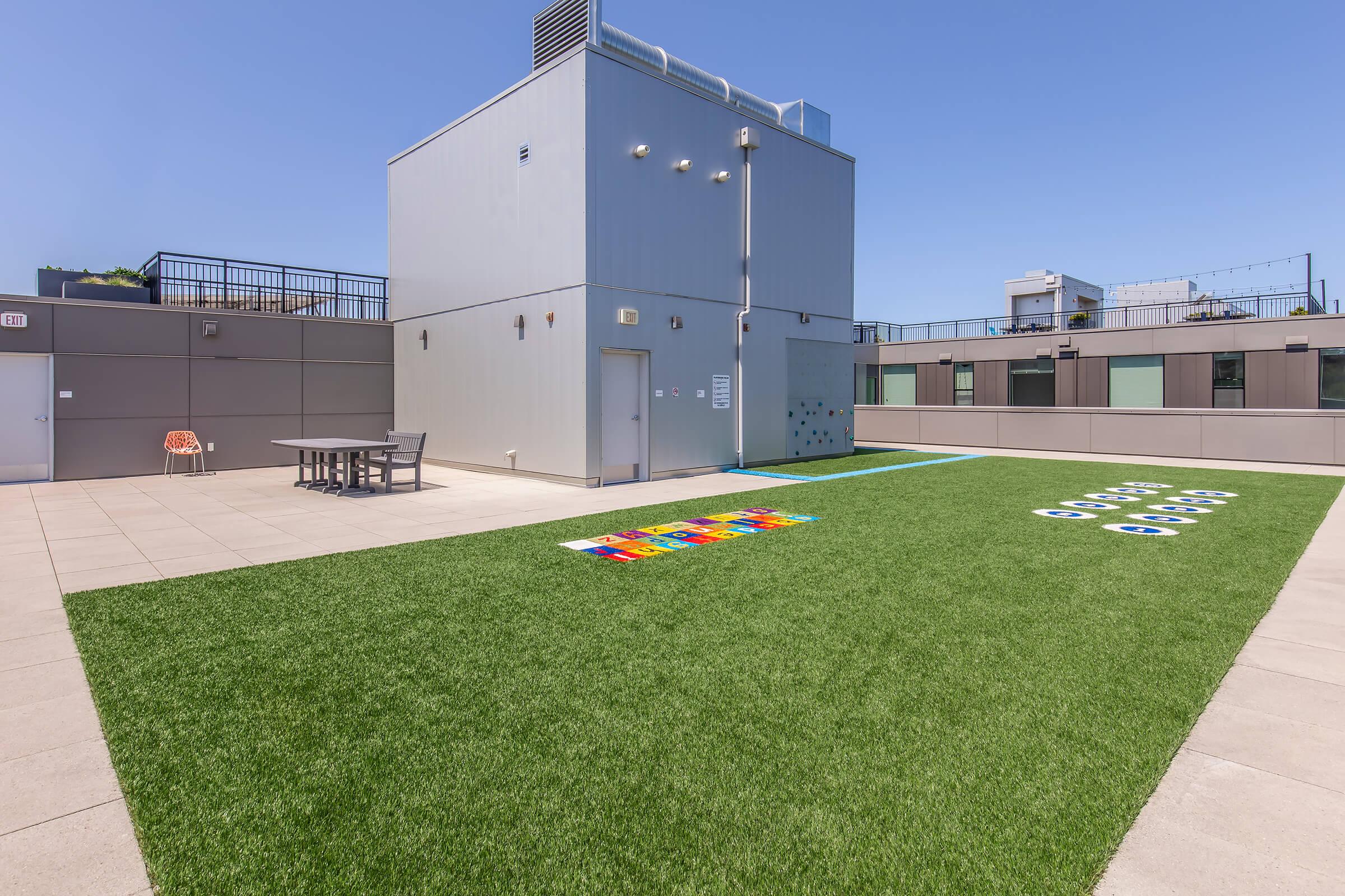 Rooftop space featuring artificial turf, a picnic table, and colorful outdoor games. The area is sunny and includes a gray building structure in the background with minimal furnishings, perfect for relaxation or social activities.