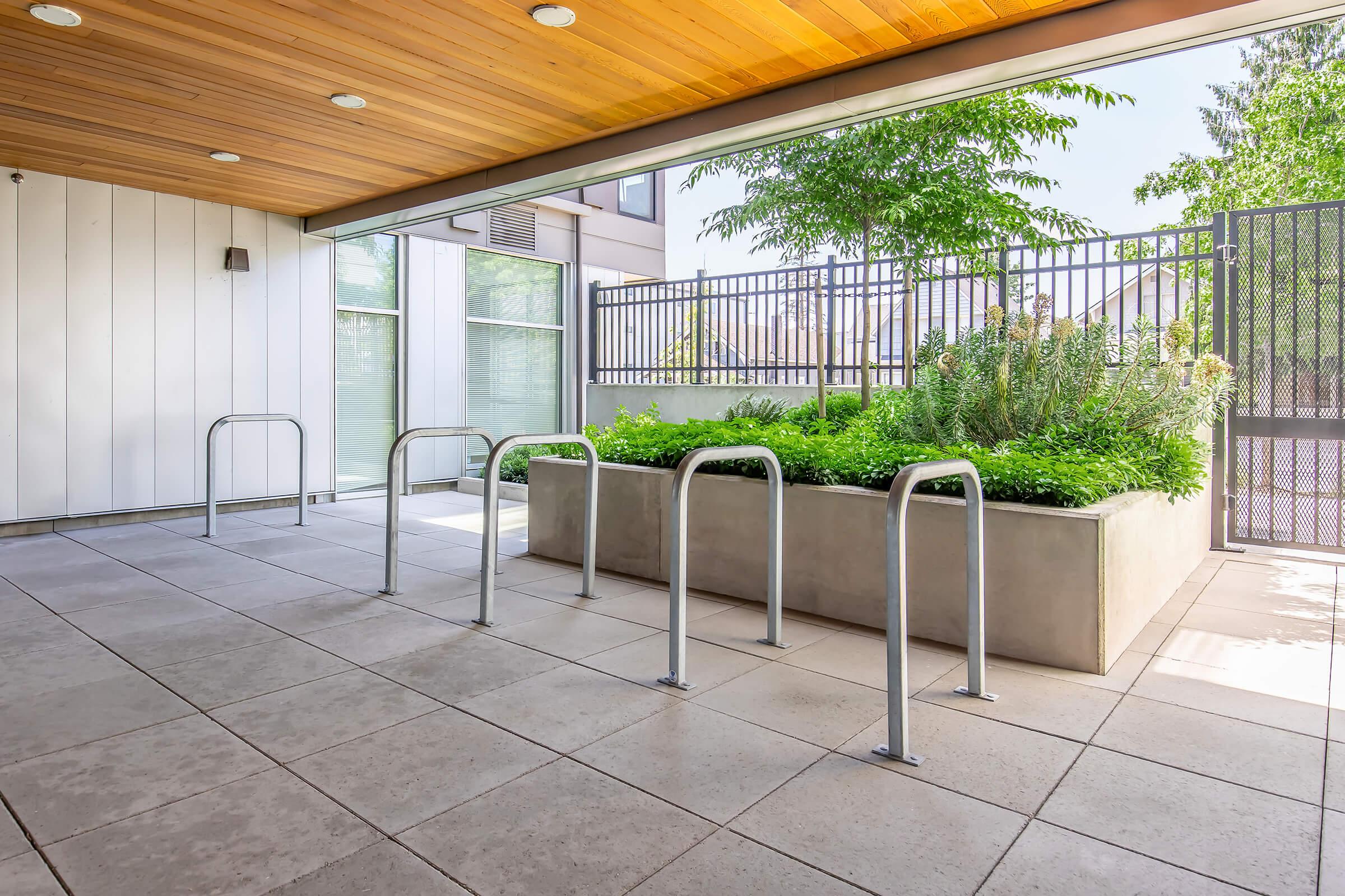 A modern outdoor area featuring a concrete planter with greenery, surrounded by a metal fence. The space includes several bike racks in the foreground, with a ceiling of wooden paneling and large windows in the background, allowing natural light to enter.