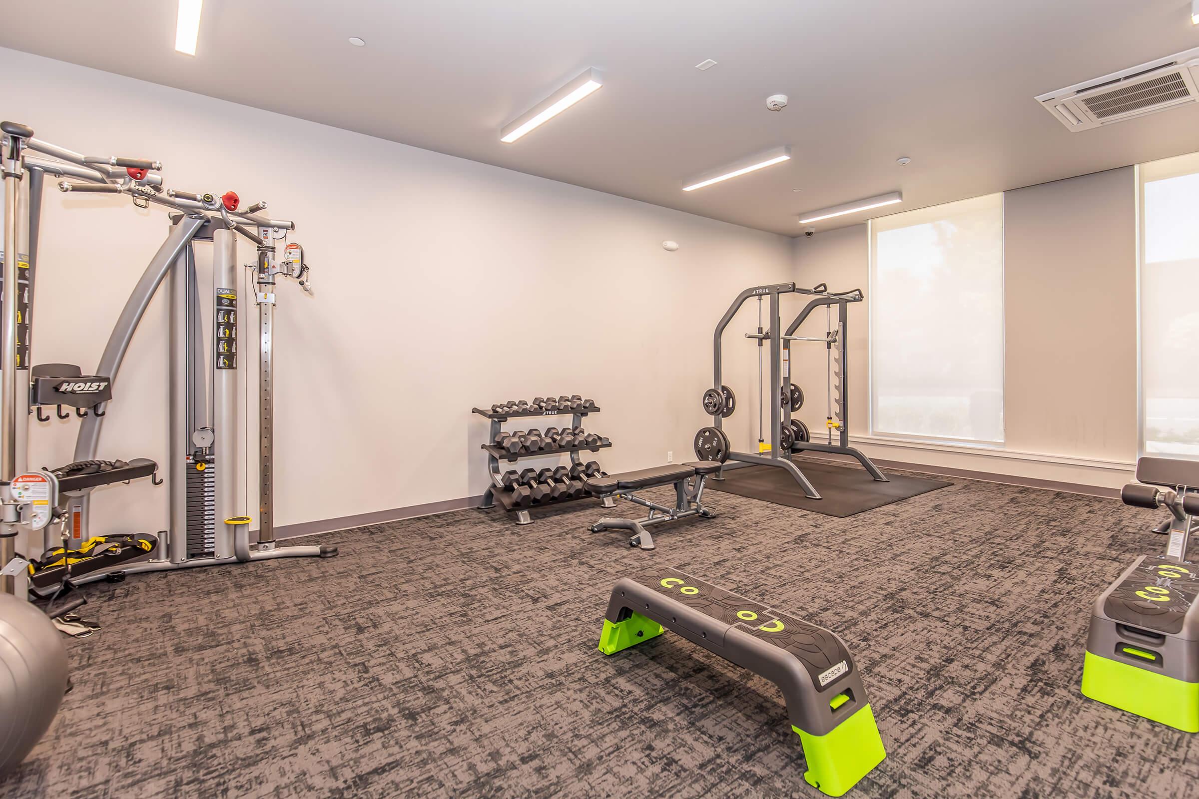 A modern gym interior featuring various exercise equipment, including weightlifting machines, dumbbells on a rack, and workout benches. The space has grey walls, bright overhead lighting, and large windows allowing natural light. The floor is covered with dark, textured carpeting.