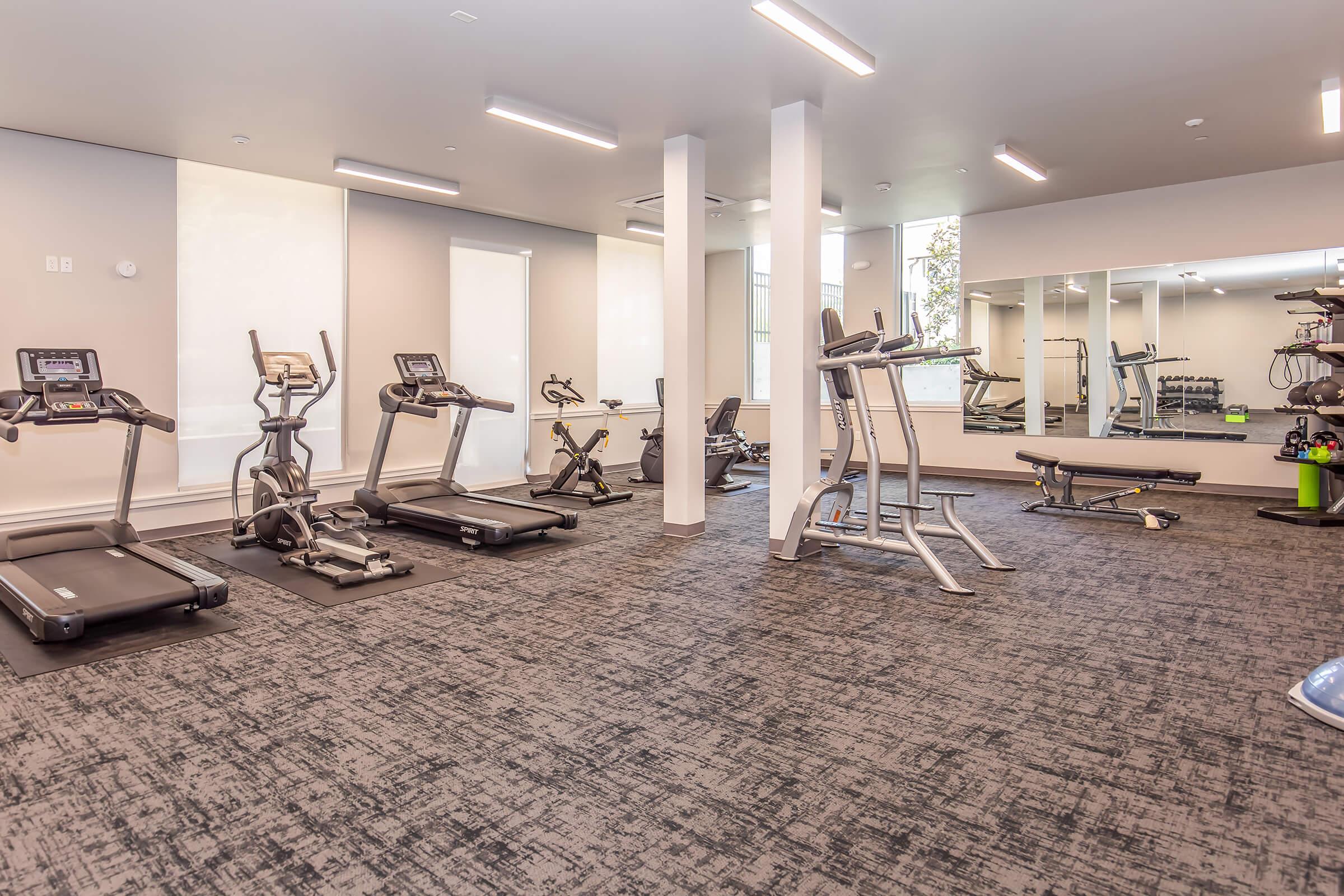 A modern fitness gym featuring several exercise machines including treadmills, elliptical trainers, and stationary bikes. The room is well-lit with large mirrors and a floor covered in dark carpeting, creating a clean and inviting workout environment.