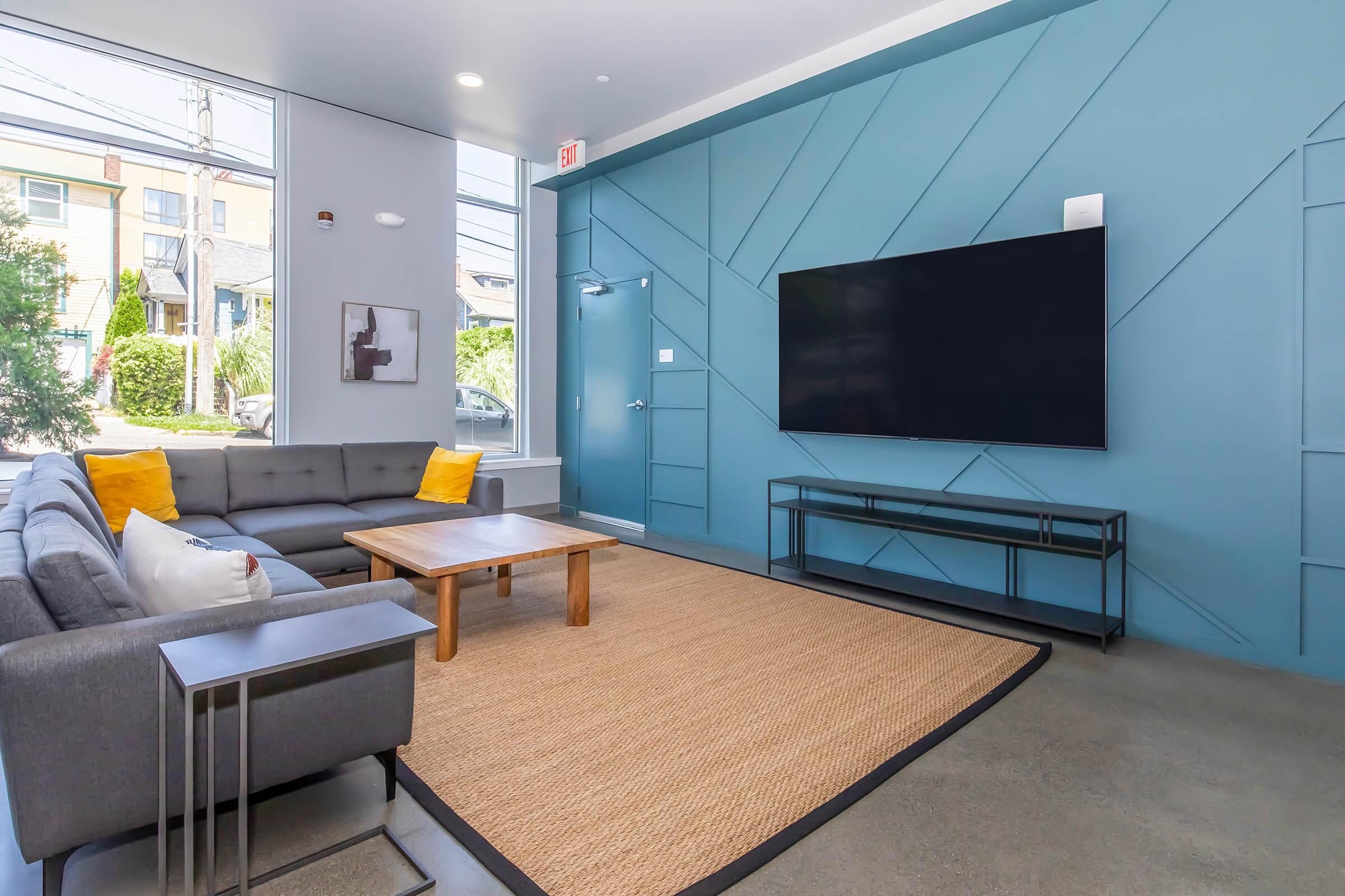 A modern living room featuring a gray sofa with yellow accent pillows, a wooden coffee table, and a large flat-screen TV mounted on a blue accent wall. There's a woven rug on the floor and sunlight streaming in through a large window, creating a bright, inviting atmosphere.