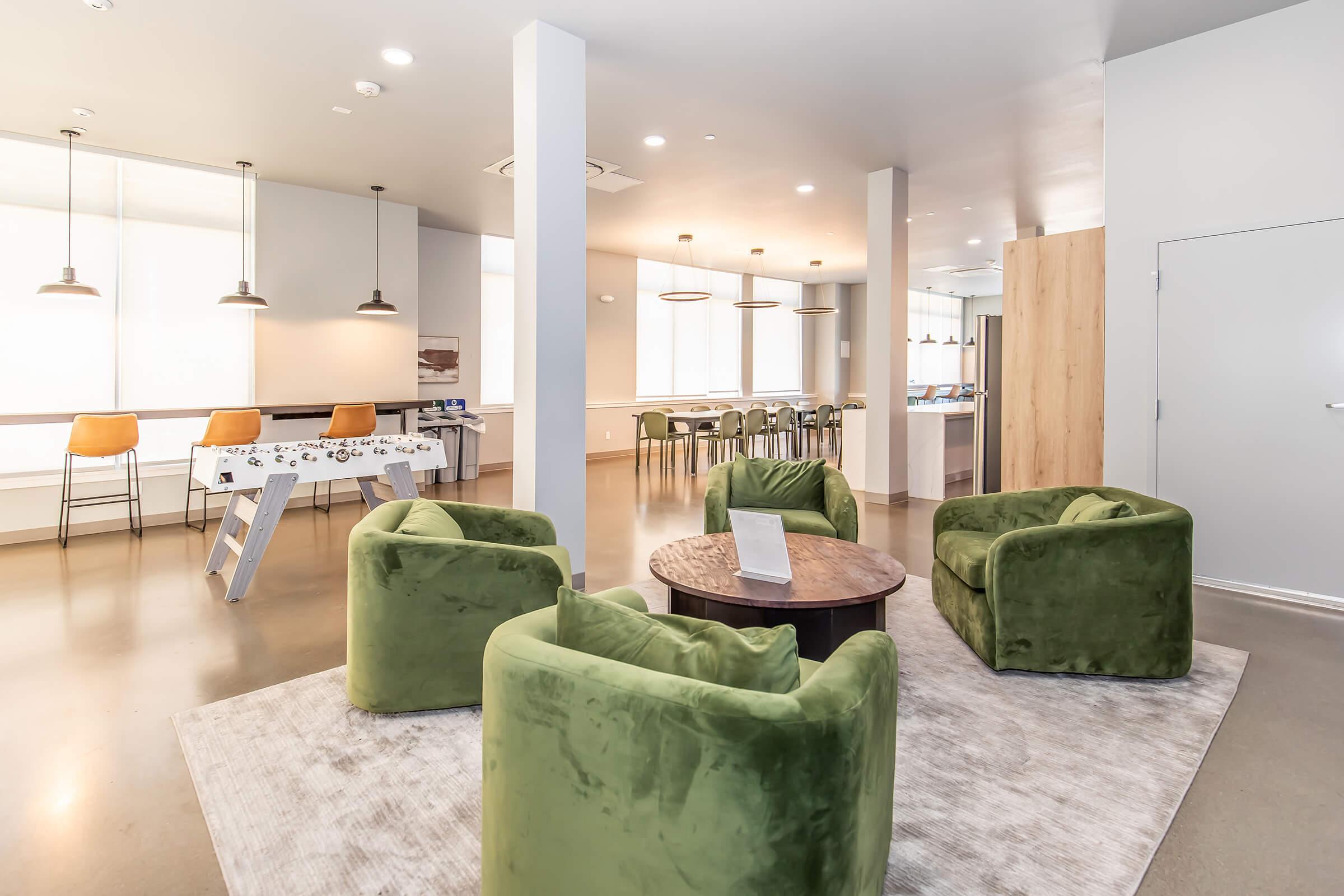 A modern lounge area featuring four green velvet armchairs arranged around a circular wooden table. In the background, there is a bar-style seating area with high stools, and large windows provide natural light, creating a bright and inviting atmosphere. The floor is polished concrete and a light rug adds warmth.