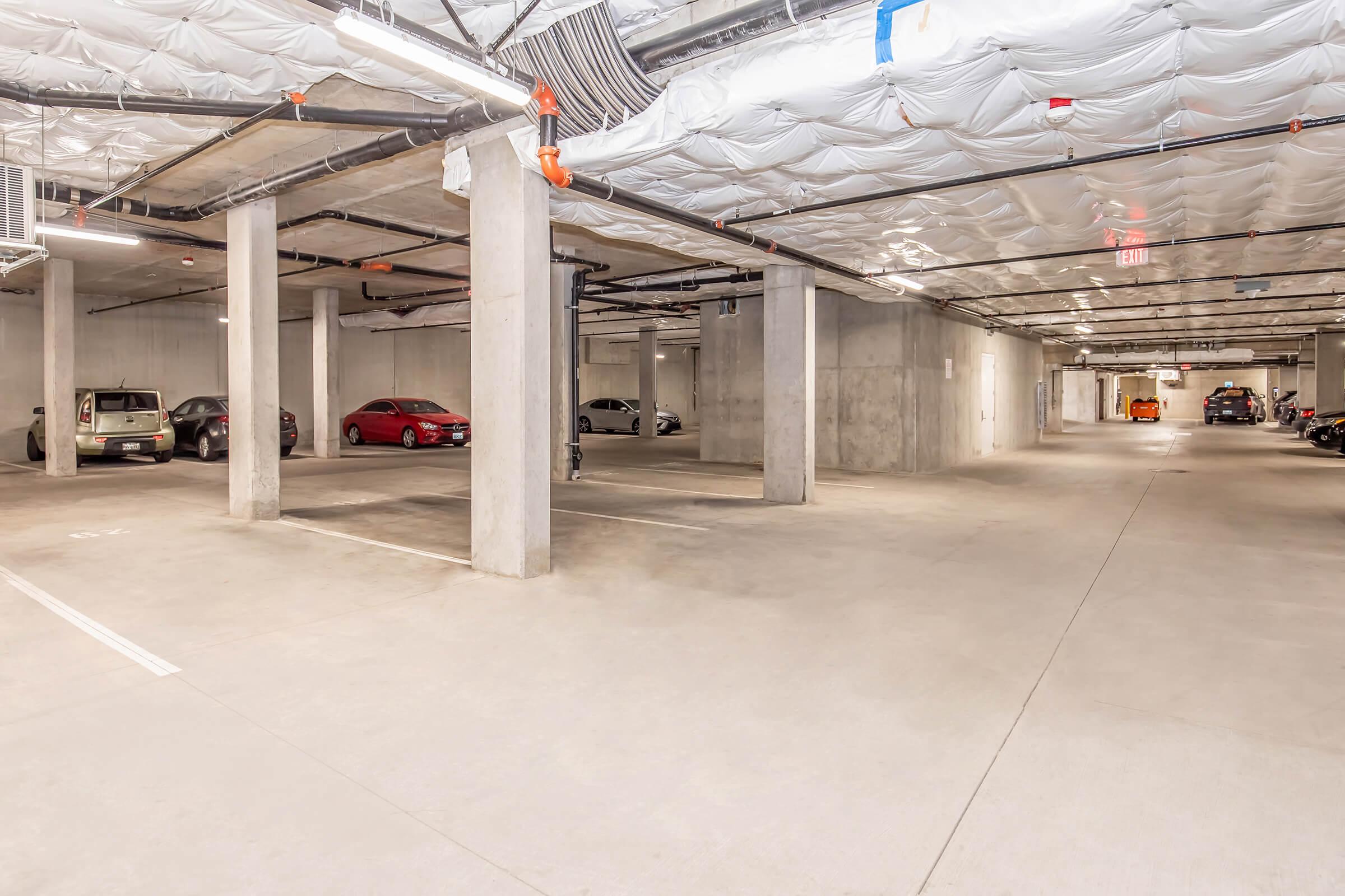 A spacious, underground parking garage featuring concrete pillars, low ceilings with insulation, and a mix of parked vehicles, including sedans and a motorcycle. The area is well-lit with visible exit signs, and the floors have clear parking lines.