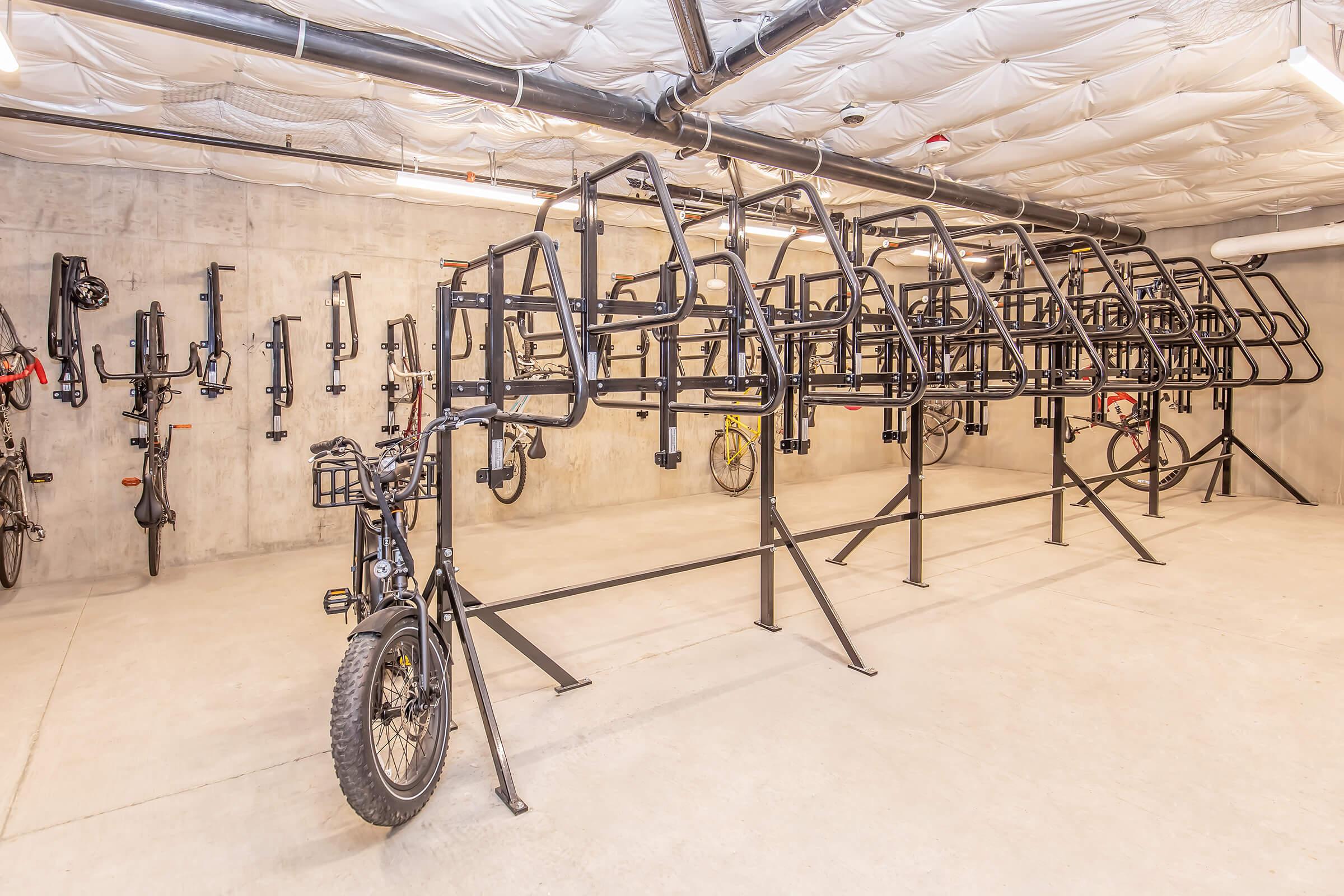 A spacious indoor bike storage area featuring multiple bike racks. Several bicycles are securely mounted on the racks, while additional bike frames are stored on the wall. The concrete floor and ceiling are visible, along with overhead lighting. The environment appears clean and organized, suitable for bike enthusiasts.