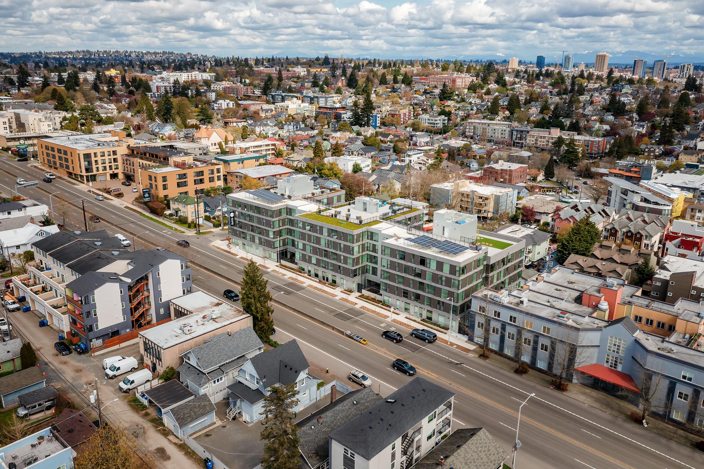 Aerial view of a suburban area featuring a mix of residential buildings and businesses. In the foreground, a modern multi-story building with green roofing is adjacent to a busy street. Surrounding the area are diverse homes and commercial structures, with patches of greenery and a backdrop of hills and cloudy skies.