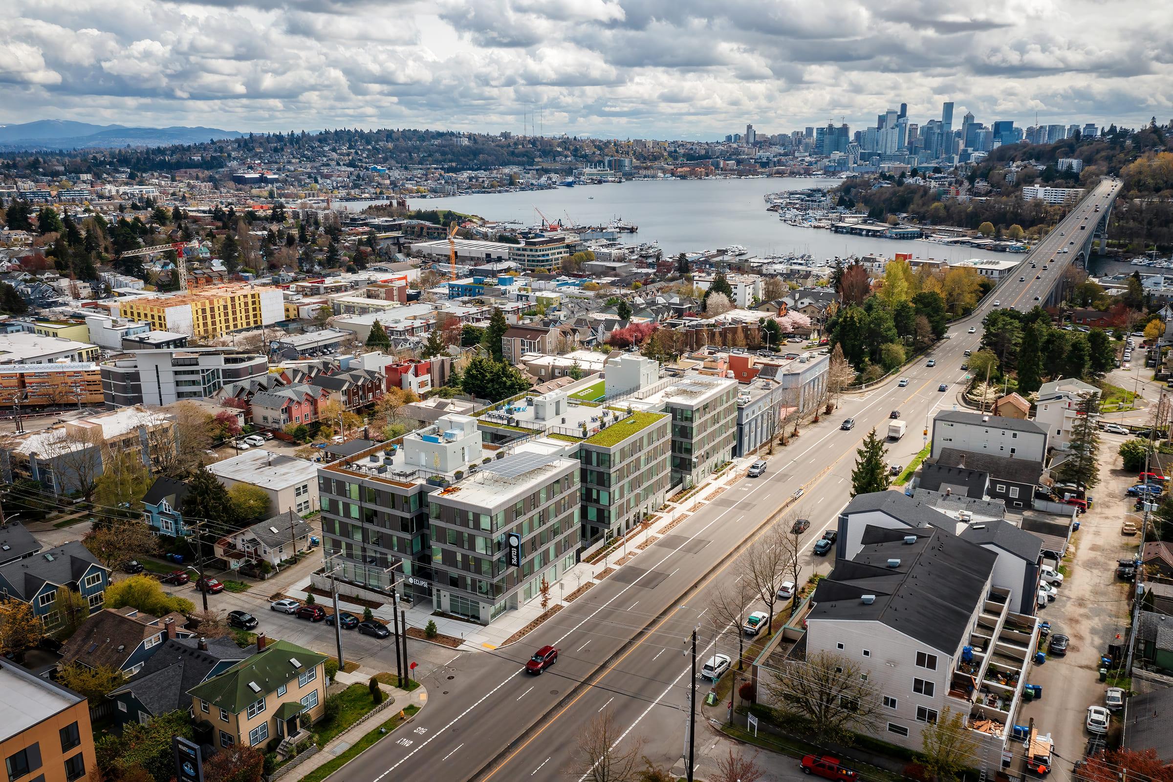 Aerial view of a vibrant Seattle neighborhood featuring modern buildings, green rooftops, and a scenic waterfront. In the background, the Seattle skyline is visible with a mix of residential and commercial areas, while a highway runs along the foreground, showcasing urban life and transportation.