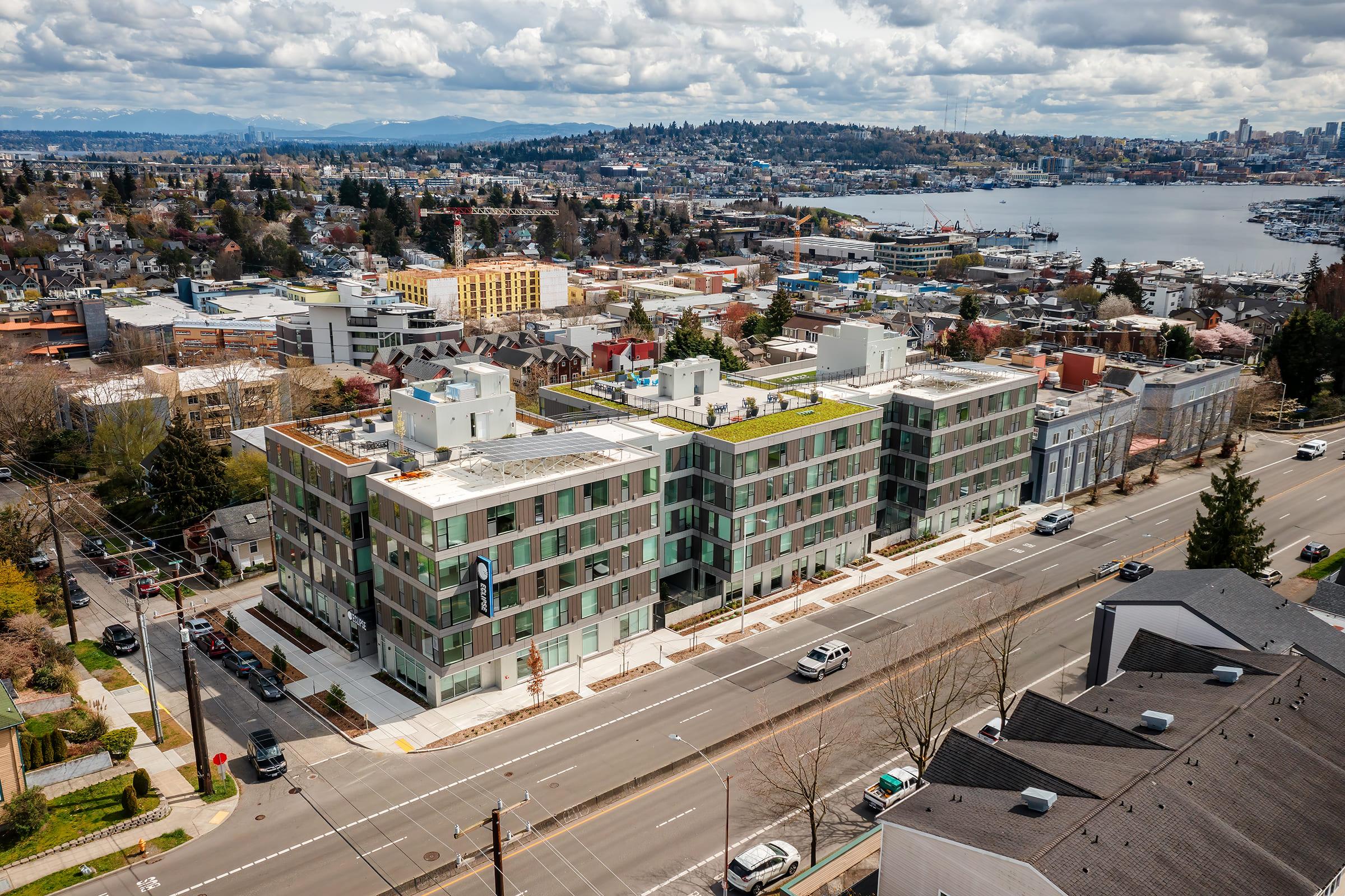 Aerial view of modern apartment buildings along a busy street, with green rooftops and large windows. The background features a scenic waterfront and a city skyline under a partly cloudy sky. Additional buildings and trees are visible, showcasing an urban landscape.