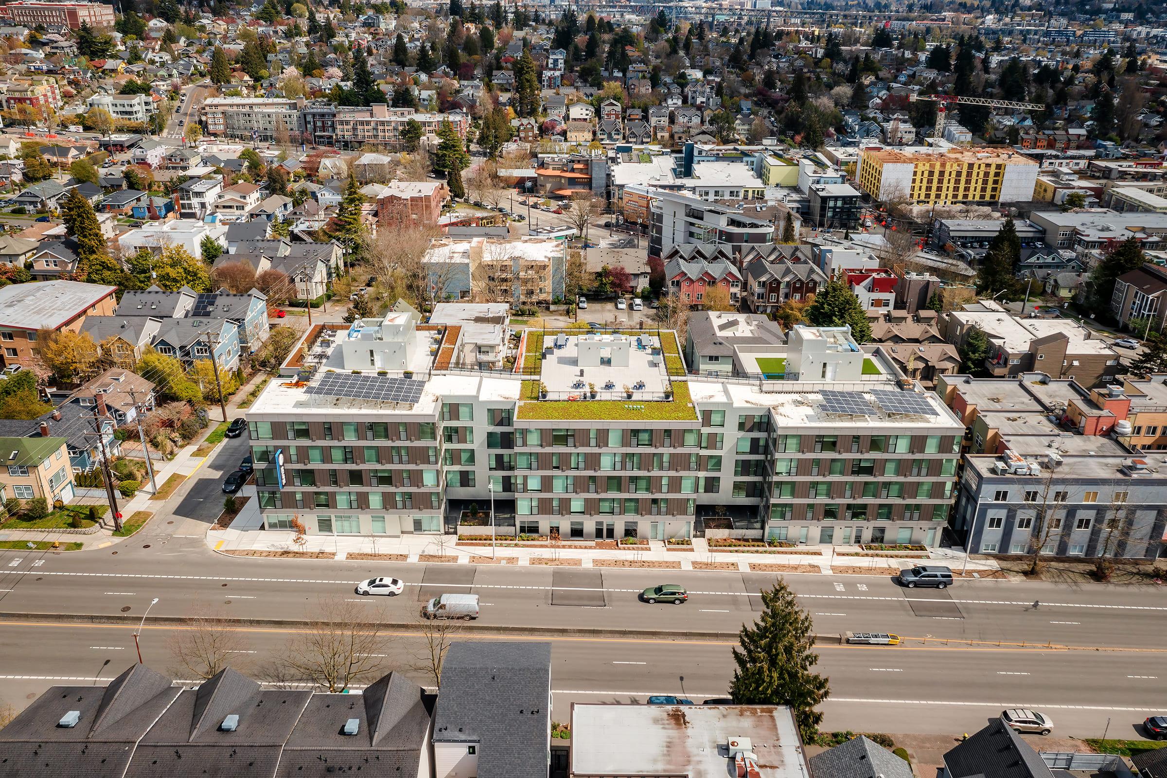 Aerial view of a modern multi-story building with a green roof, surrounded by a neighborhood of homes and trees. The street in front features parked cars and a busy thoroughfare, while the background shows a mix of residential and commercial buildings, along with a glimpse of distant hills.