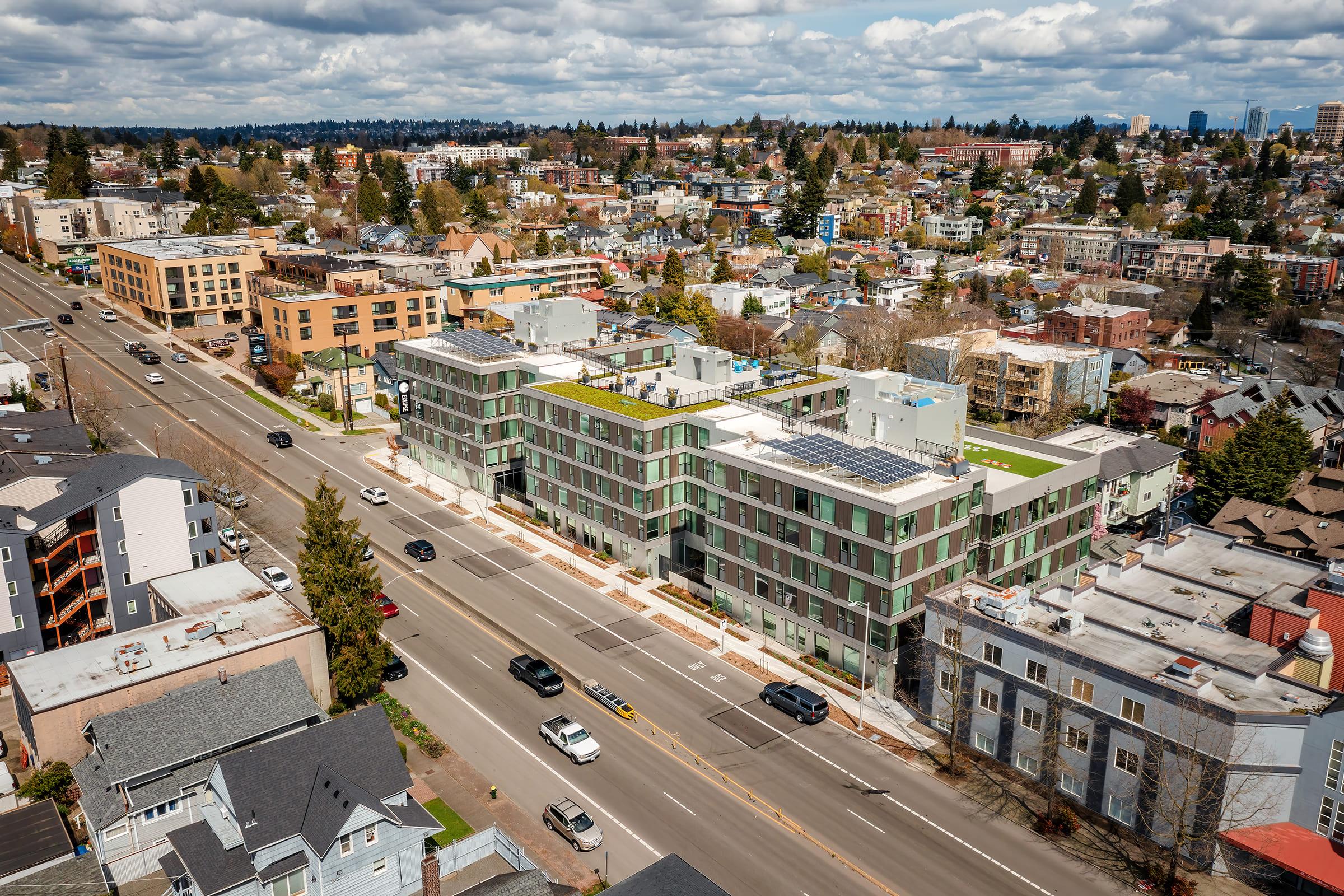 An aerial view of a modern, multi-story residential building with green rooftops, surrounded by a mix of urban development, including commercial buildings and houses. The street in the foreground shows cars driving by, and the sky is partly cloudy, indicating a clear day.