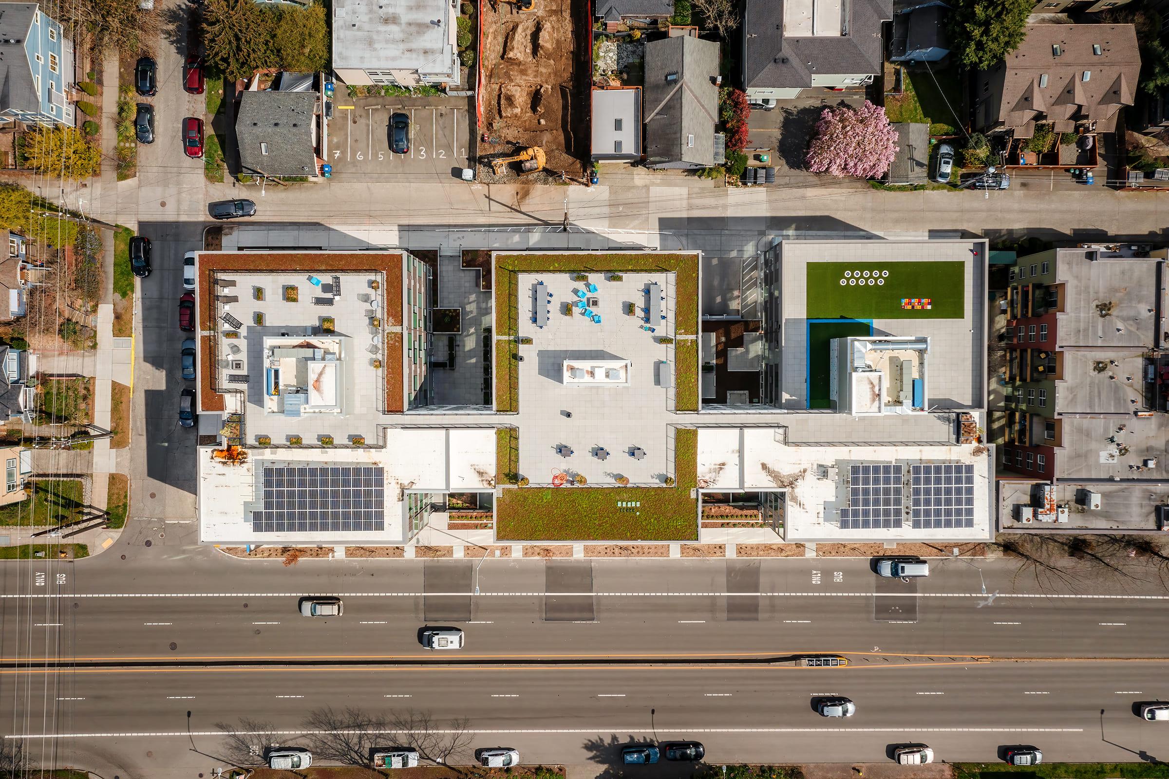 Aerial view of a modern urban complex featuring green rooftops and solar panels. Surrounding buildings are visible, along with a road filled with cars. The rooftop areas include open spaces with seating and recreational facilities, indicating a blend of residential and communal spaces.