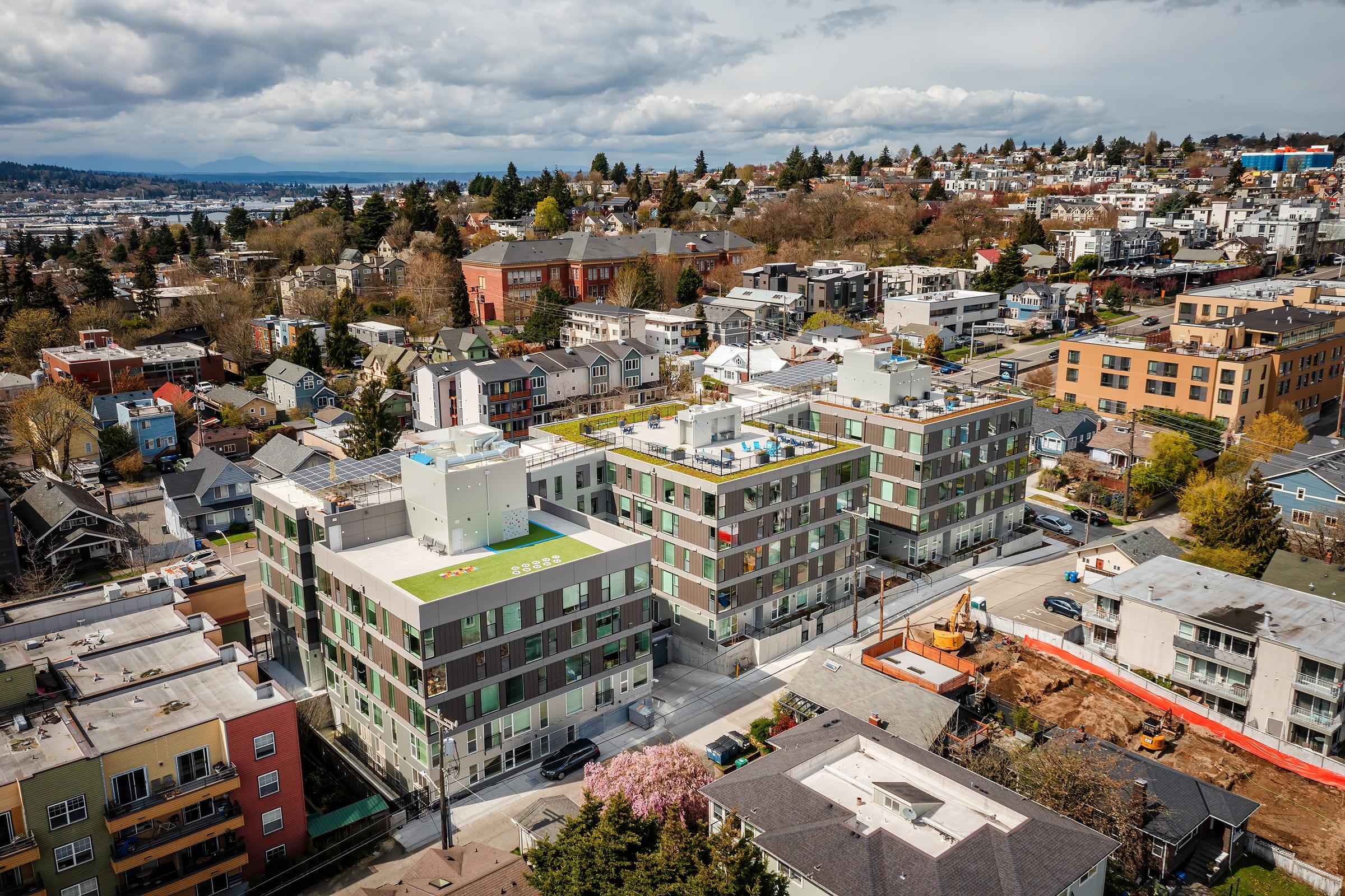 Aerial view of a residential neighborhood featuring several multi-story apartment buildings surrounded by smaller homes. The scene includes trees, parked cars, and construction activity, with a backdrop of cloudy skies and distant hills.